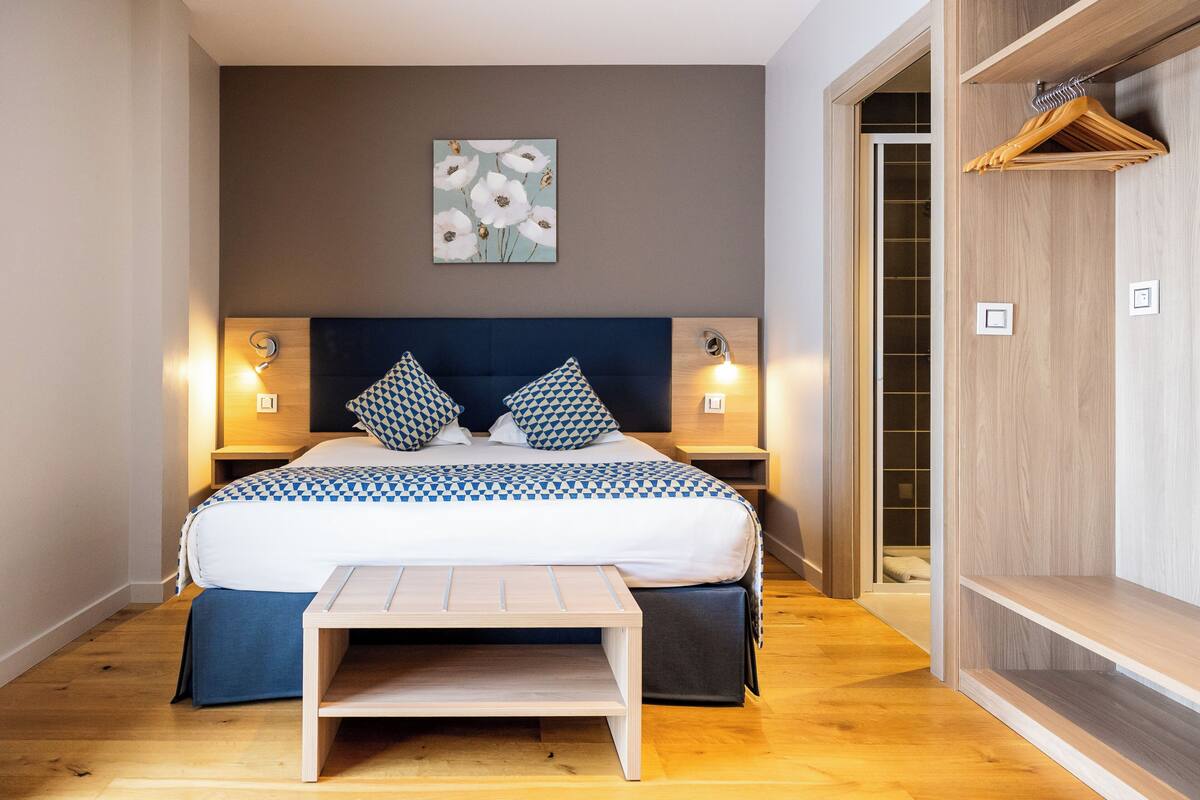 A bedroom with a bed, bedside tables, a bench, a wooden wardrobe, and a wall-mounted artwork.