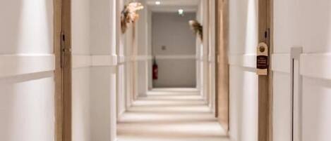 A long, narrow hallway with white walls and doors on both sides, leading to a room at the end.