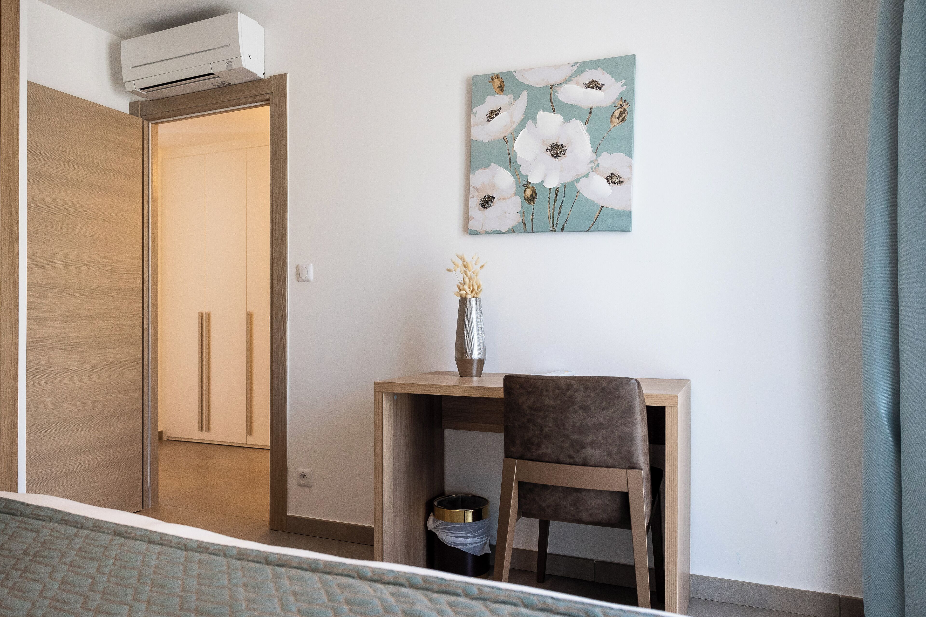 Ein Hotelzimmer mit Bett, Schreibtisch mit Stuhl, Blumen in einer Vase, einem Papierkorb und einem Gemälde an der Wand.