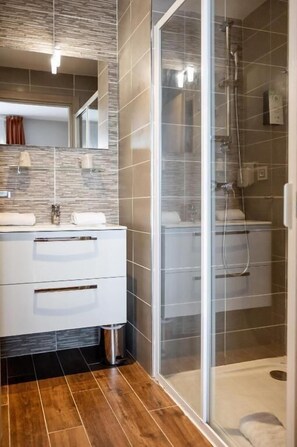 A modern bathroom with a glass shower enclosure, a white vanity with a mirror, and a wooden floor. - Hôtel Le Bastia (Bastia)