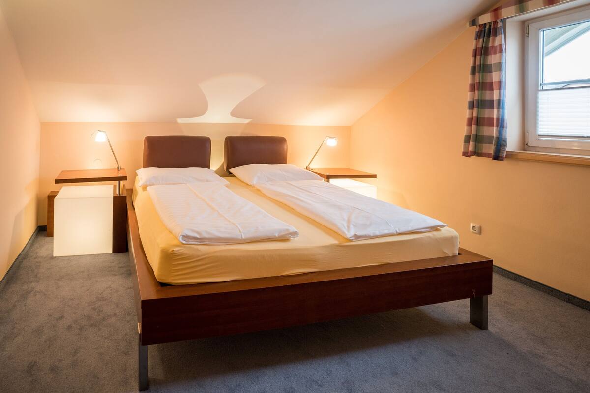 Un dormitorio con una cama, dos mesitas de noche, una lámpara, una ventana con cortinas y un aplique de pared.