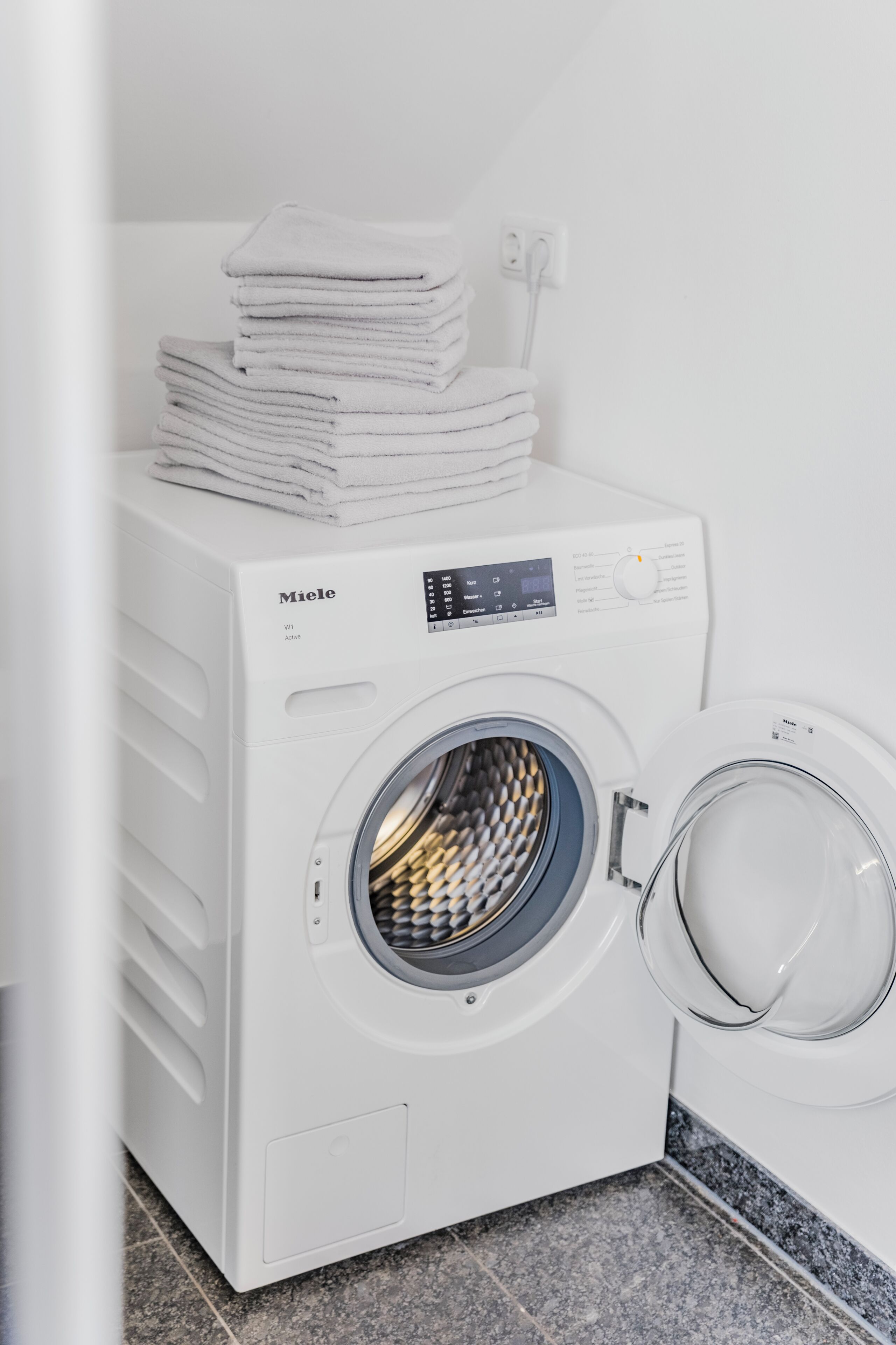 Eine weiße Miele Waschmaschine mit Bedienfeld, ein Stapel weiße Handtücher und ein gefliester Boden.