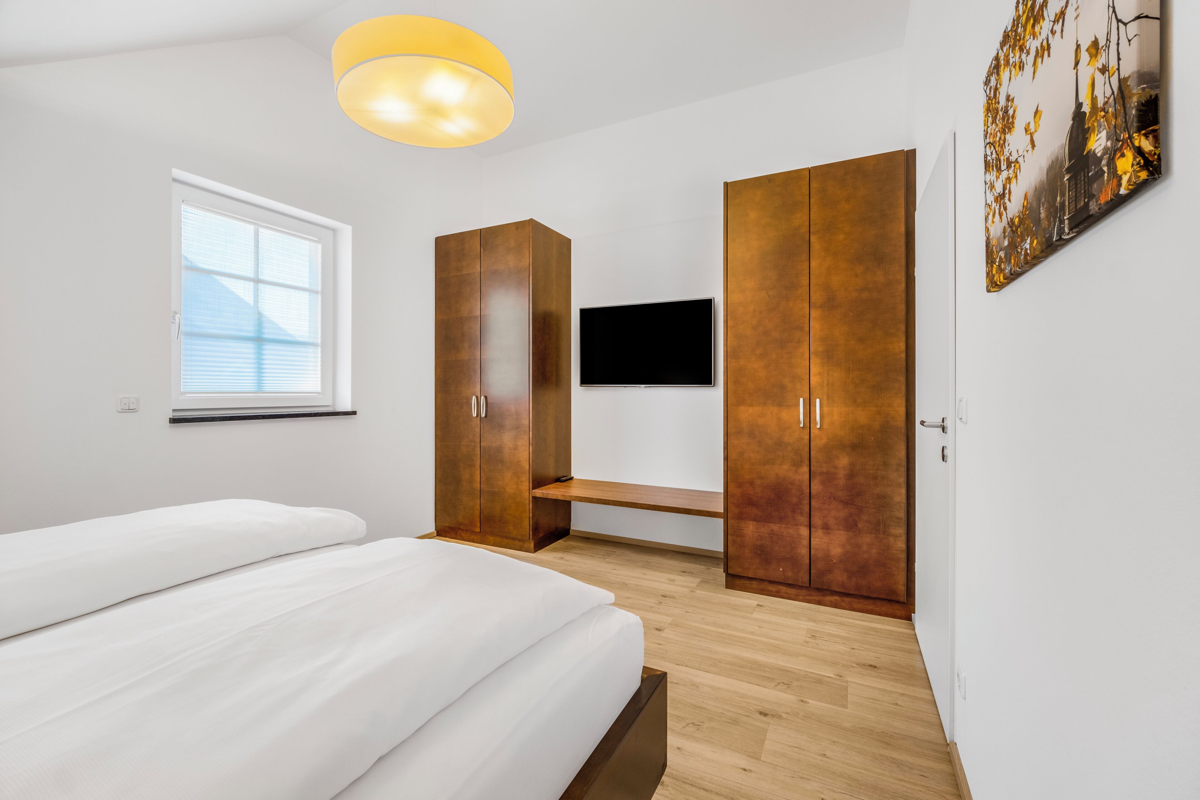 A bedroom with a bed, a TV, wooden wardrobes, a bench, and a window.