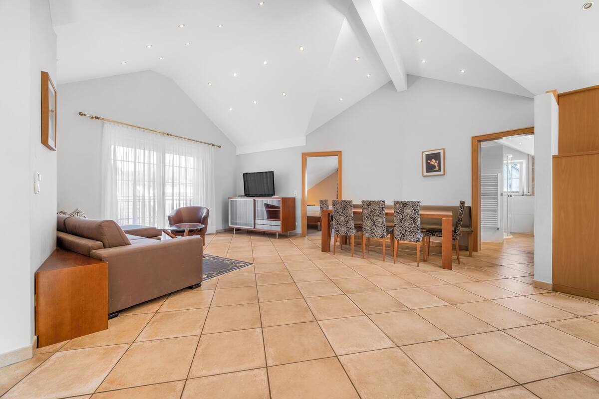 Amplia sala de estar con techo alto, sofá, televisor, mesa de comedor con sillas y vistas al exterior.