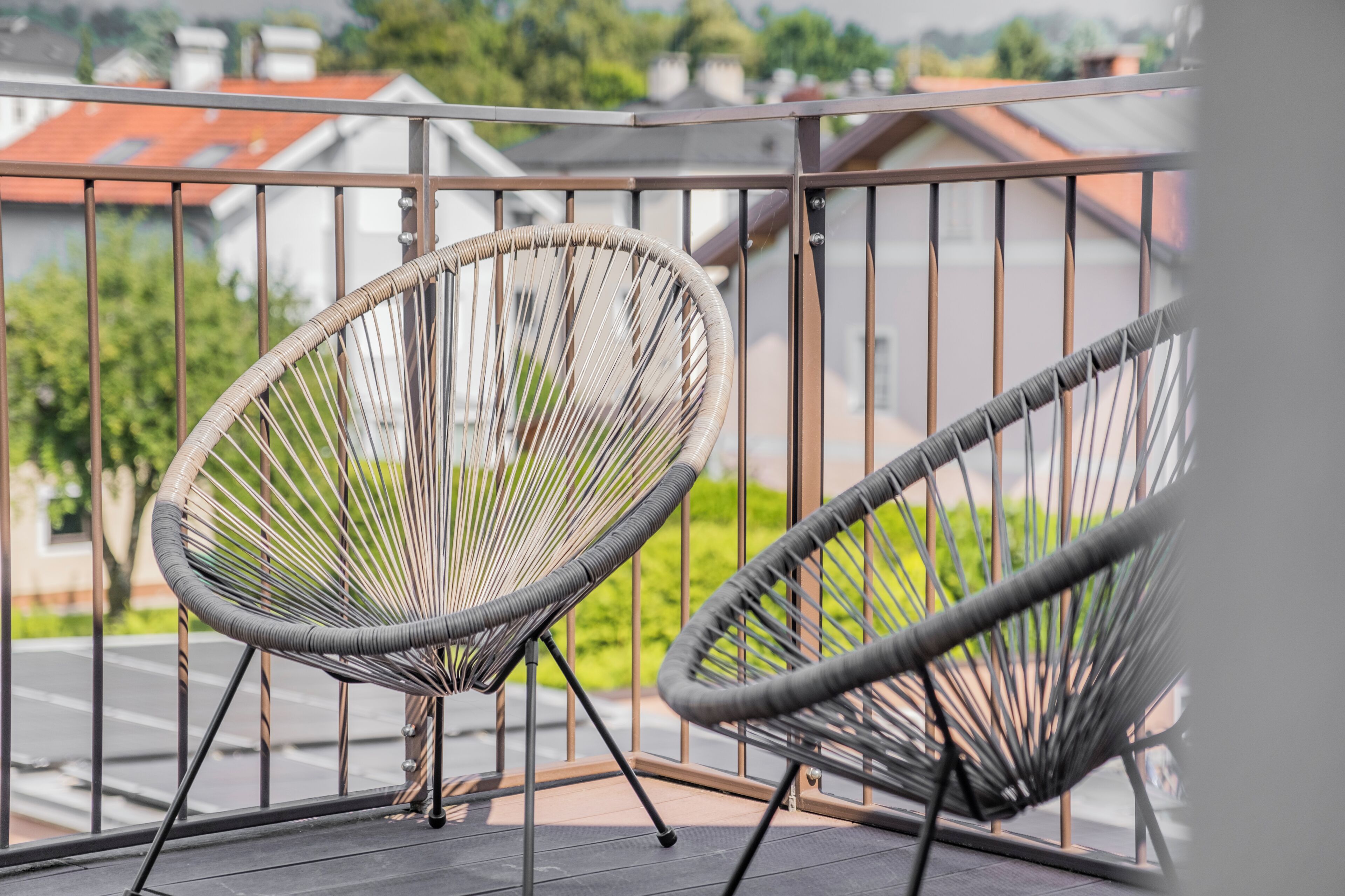 Two wicker chairs on a balcony with a metal railing, overlooking a residential area.