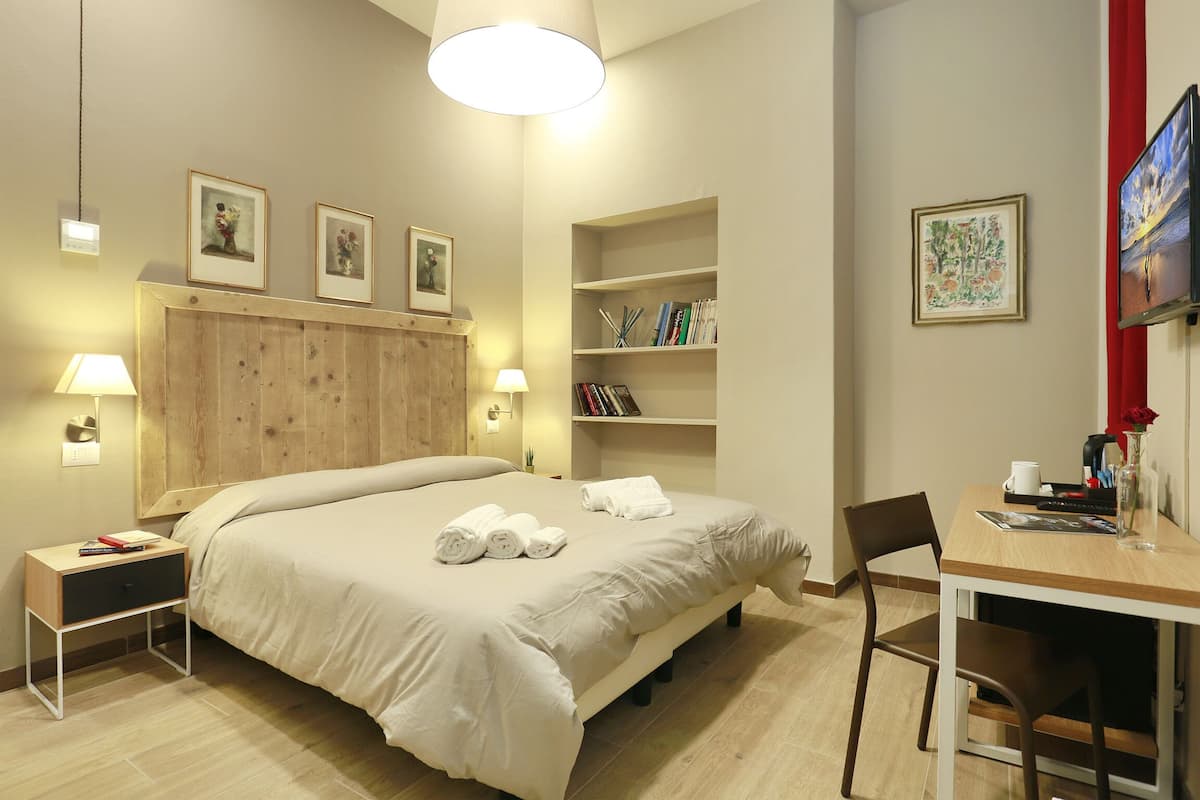 A hotel room with a bed, bedside tables, a desk, and shelves with books and decorative items.