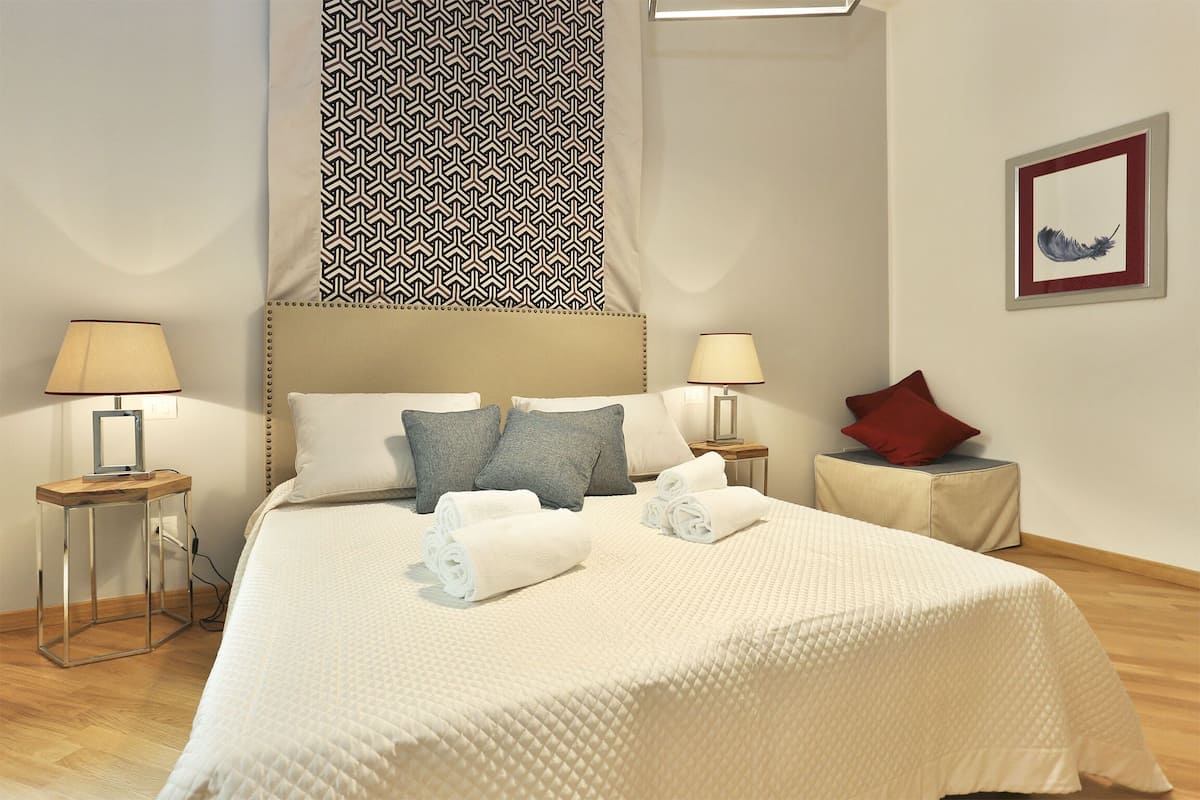 A neatly made bed with a quilted white duvet, two pillows, and rolled towels on a wooden floor. There are two bedside tables with lamps, a patterned curtain, and a framed picture on the wall.