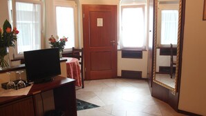 Une pièce avec une porte en bois, un téléviseur posé sur un meuble en bois, une petite table recouverte d’une nappe rouge et un miroir accroché au mur.