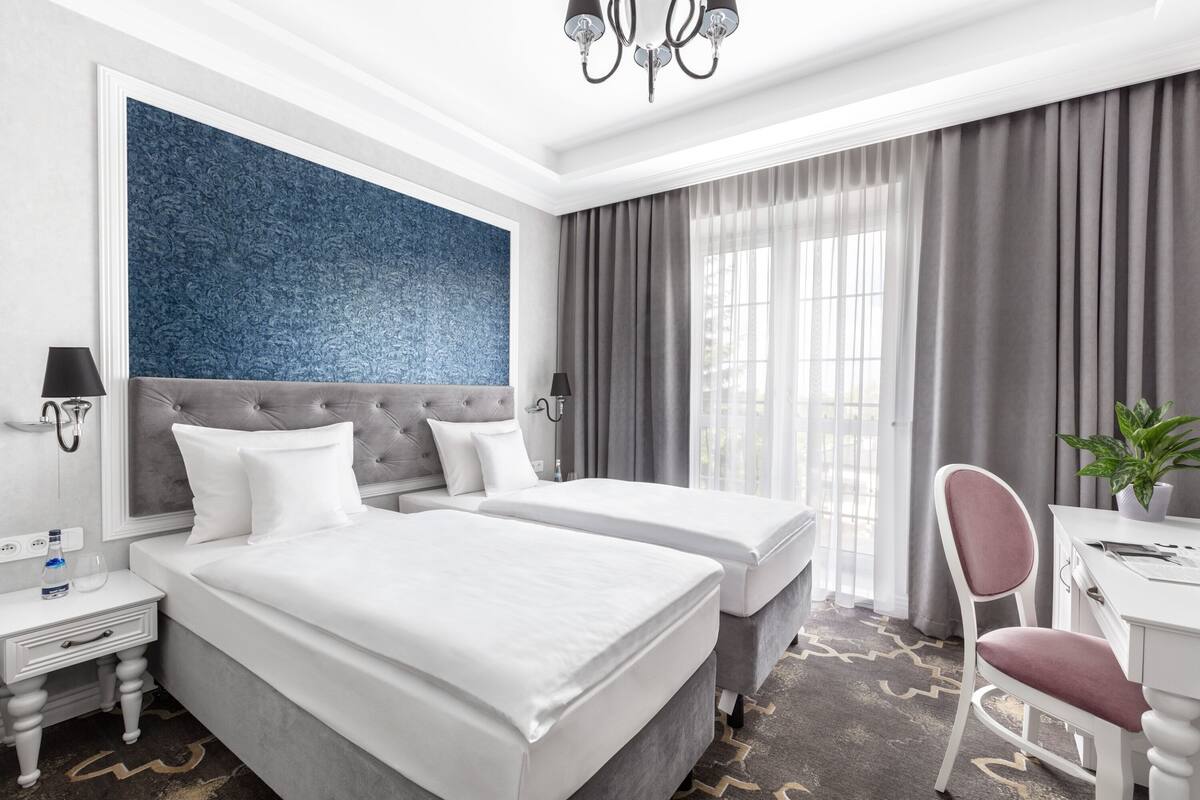 A hotel room with two beds, a desk, a chair, a potted plant, and a large blue accent wall.