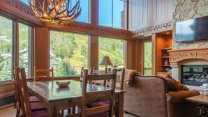 A spacious living room with a large wooden dining table, comfortable seating, a fireplace, and a television mounted on a stone wall. - 3bd + Loft With Ski In/out Access And Panoramic Views! 3 Bedroom Condo by RedAwning (Avon)
