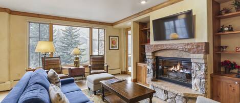 A cozy living room with a stone fireplace, a large TV mounted above it, and a comfortable seating area with a blue sofa and a beige armchair.