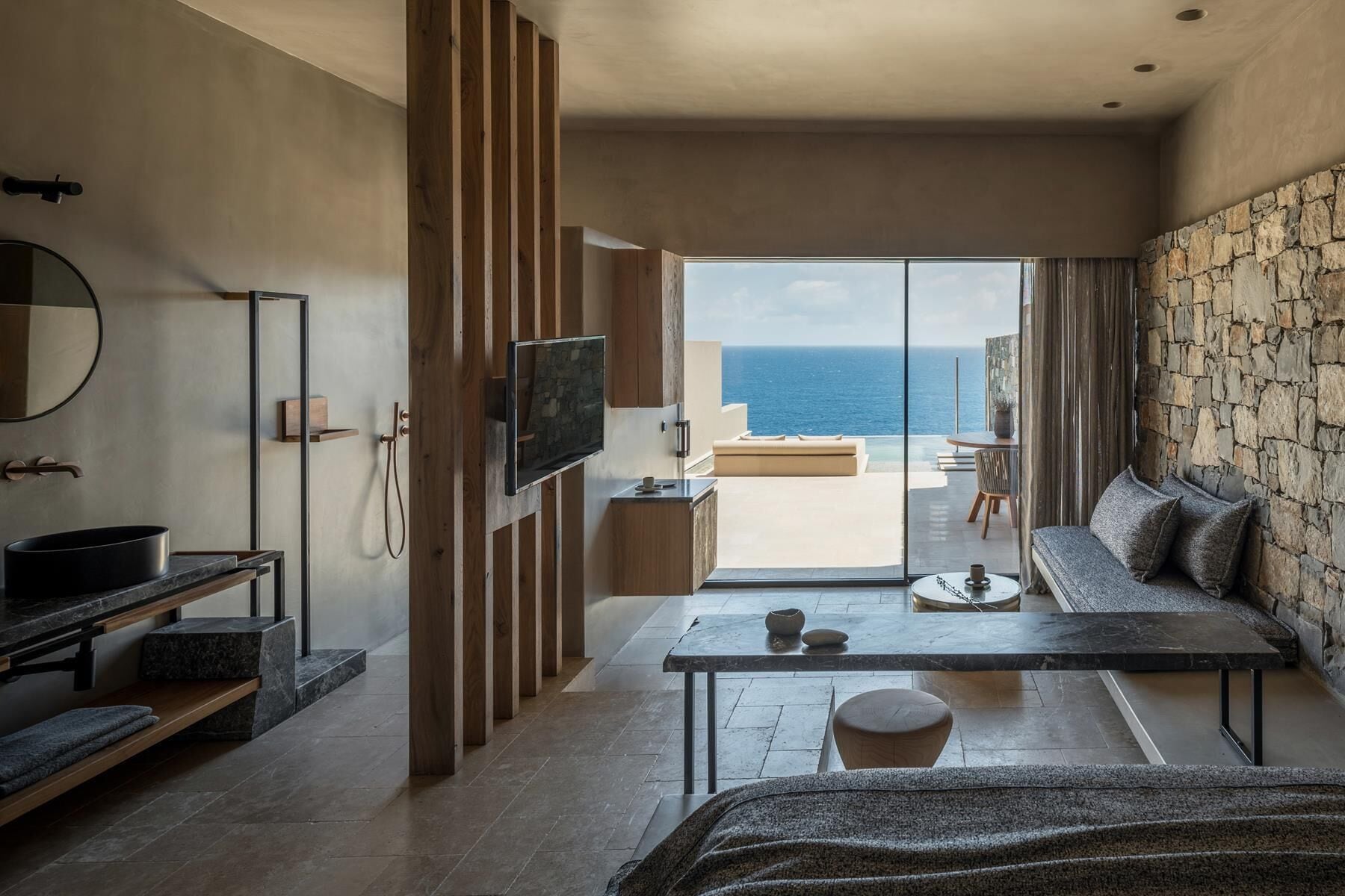 A modern hotel room with a large window offering a view of the sea, a sofa, a TV, and a minimalist bathroom.