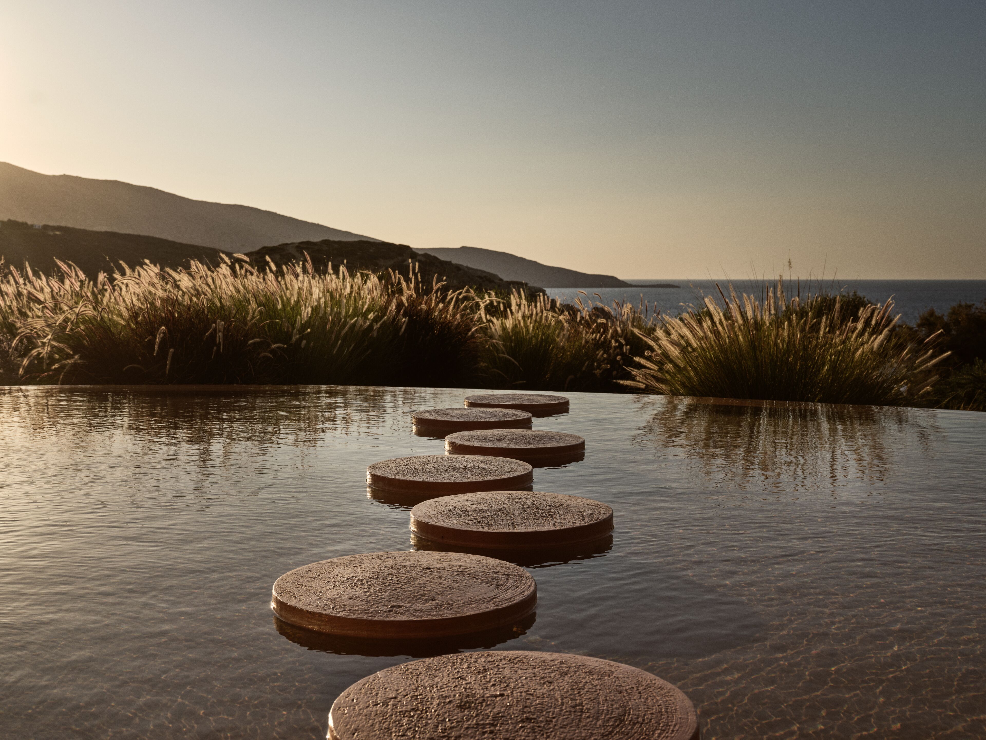 A series of circular stone steps leading into a body of water, surrounded by grass and hills in the background.
