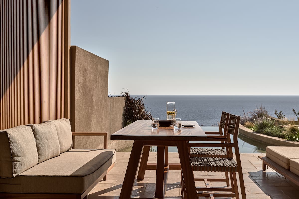 An outdoor patio with a wooden table and chairs, a beige sofa, and a view of the ocean.