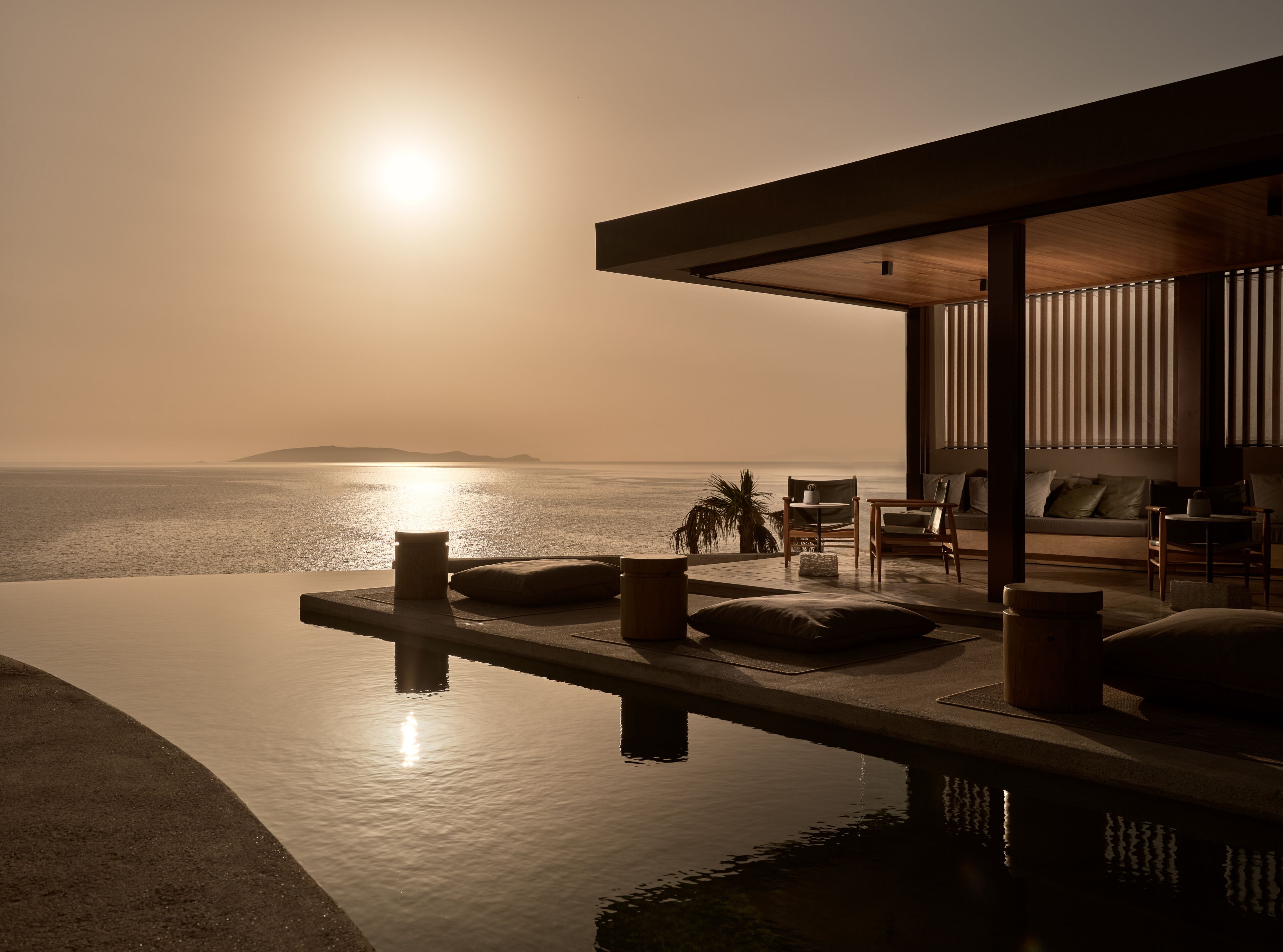 A modern infinity pool with a view of the ocean at sunset, a wooden deck, and a seating area with cushions and a table.