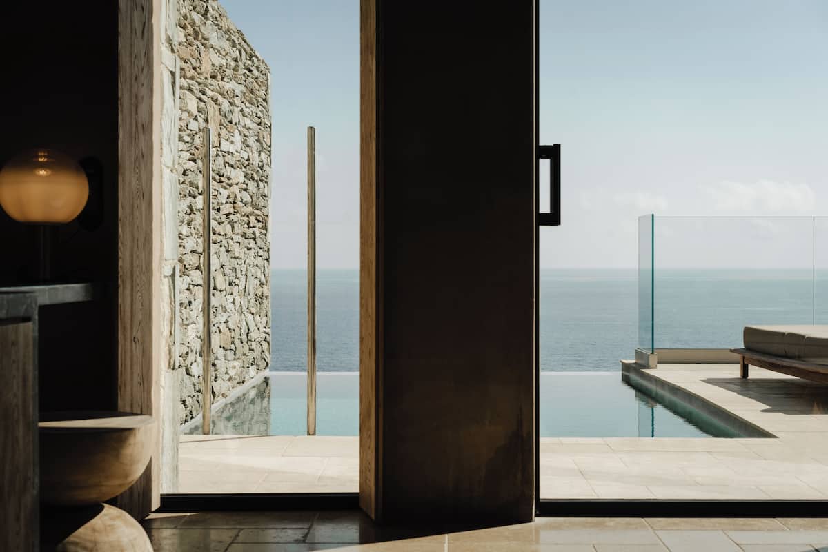 An infinity pool with a clear view of the sea, a stone wall, and a modern interior with a wooden bench and a lamp.