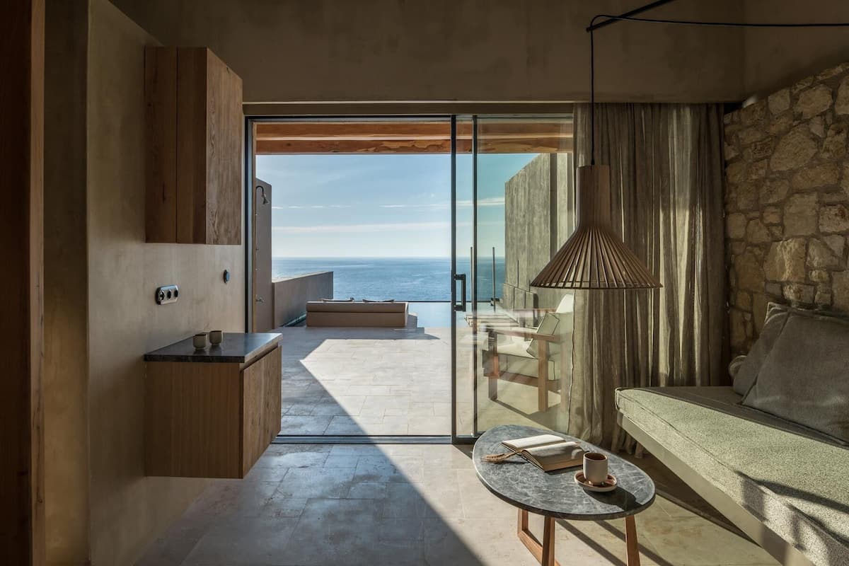 A modern interior with a stone wall, a wooden cabinet, a glass door leading to a balcony with a view of the sea, and a round table with a book and cup.