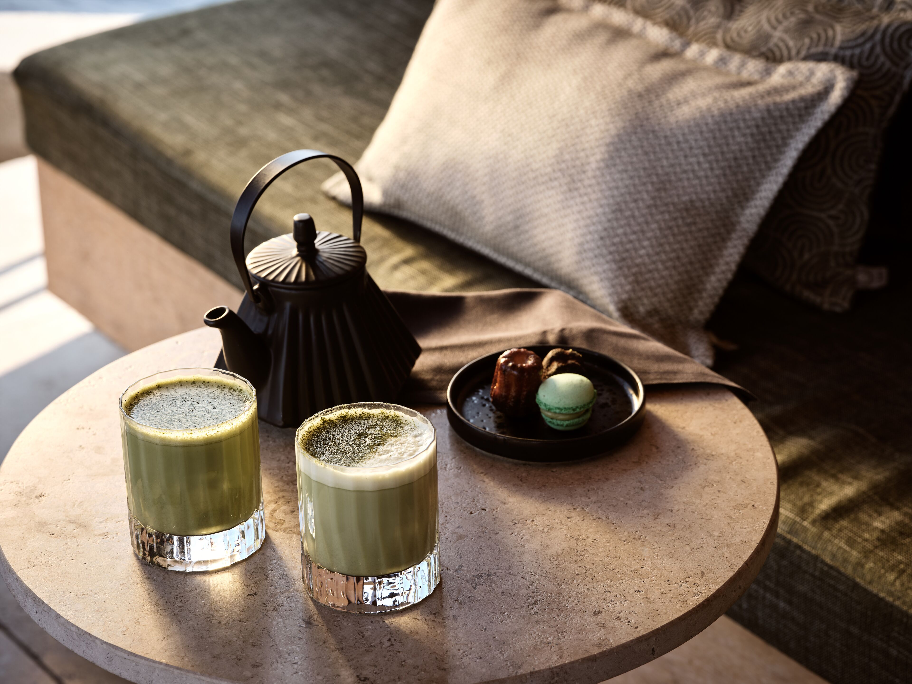 A round table with two glasses of matcha on a textured surface, a black teapot, and a plate with two macarons.