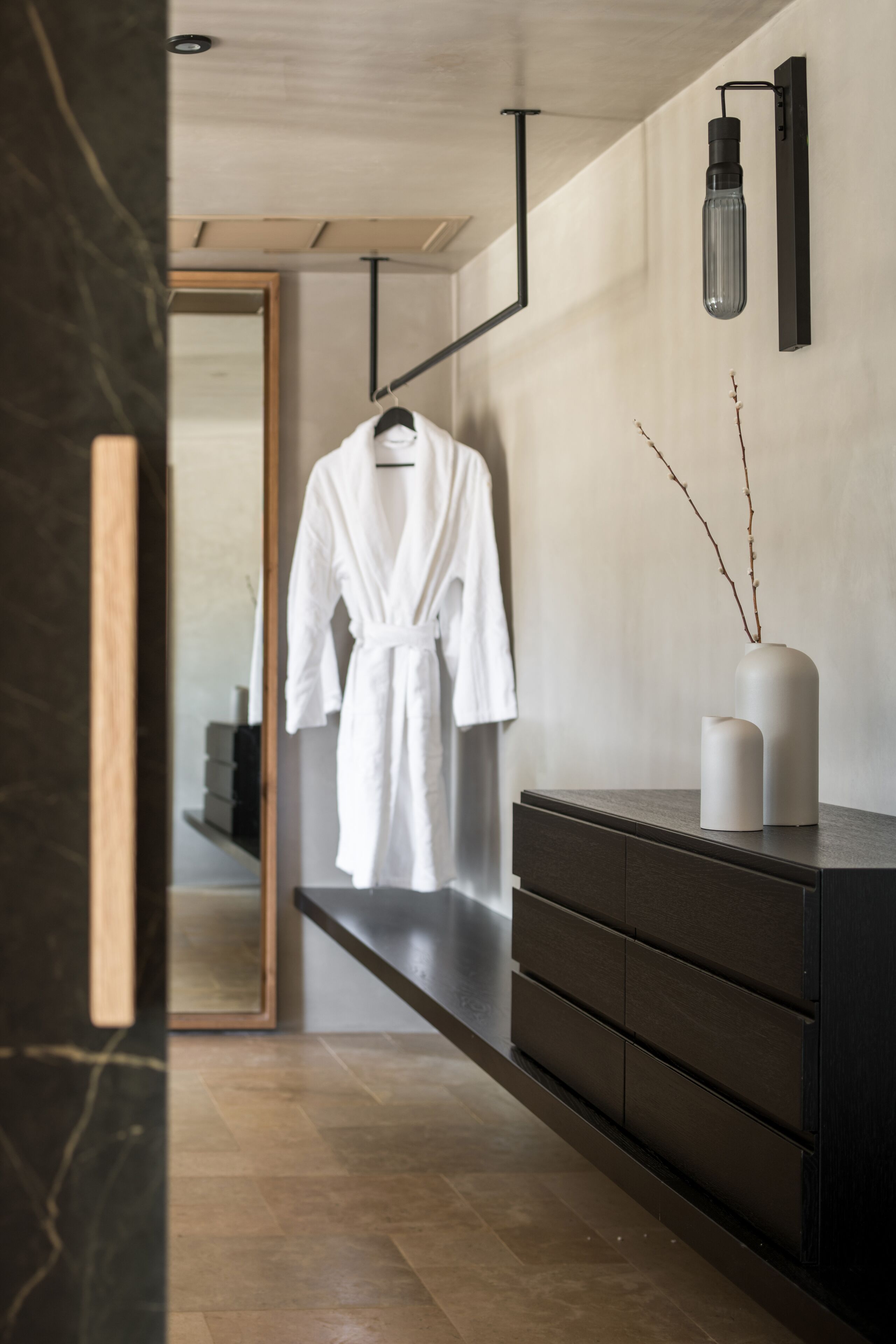 Een moderne badkamer met een houten vloer, een donker houten wastafelmeubel, een witte badjas die aan een rek hangt, en een vaas met takken op het wastafelmeubel.