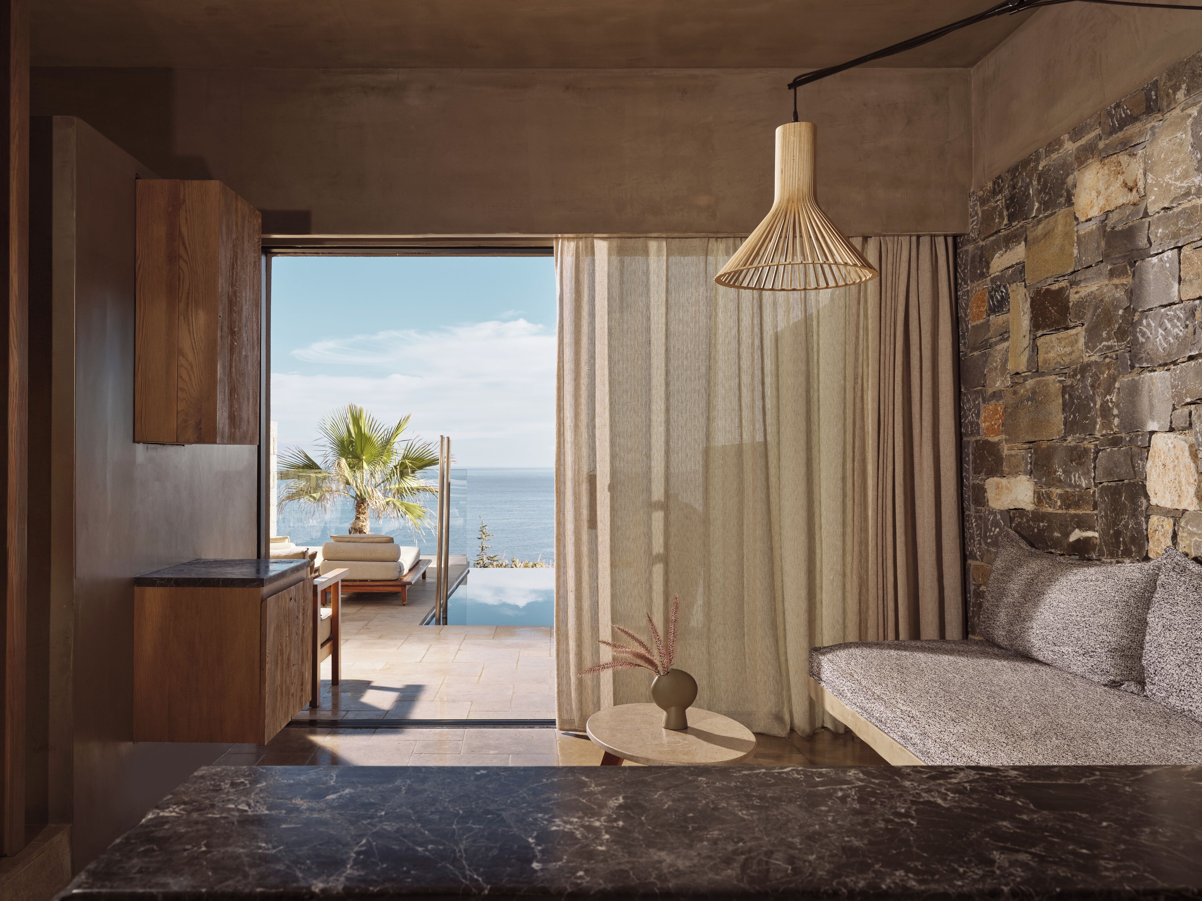 A modern room with a stone wall, a view of the ocean, a sofa, a small table with a vase, and a hanging light fixture.