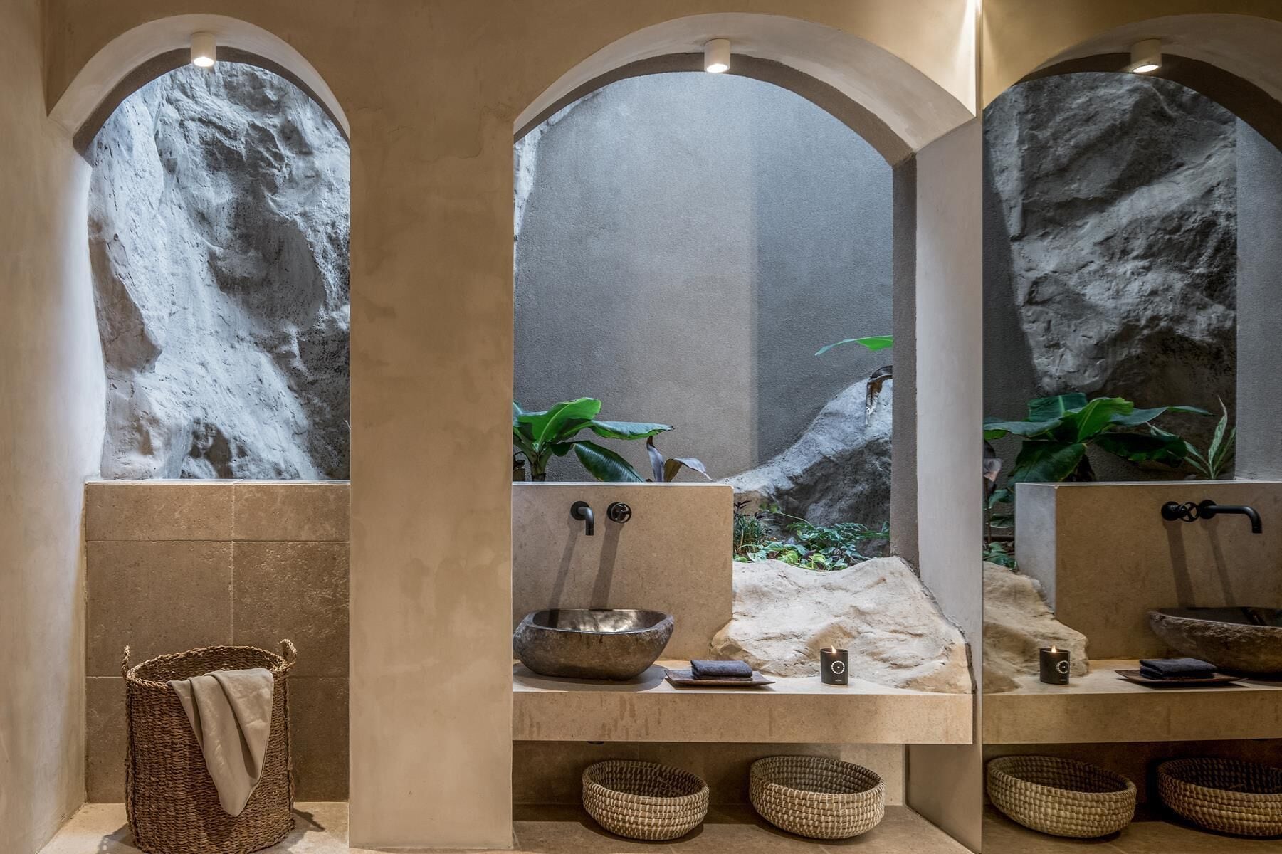 A bathroom with a central sink, a mirror, and a stone wall. There are three woven baskets on the floor.