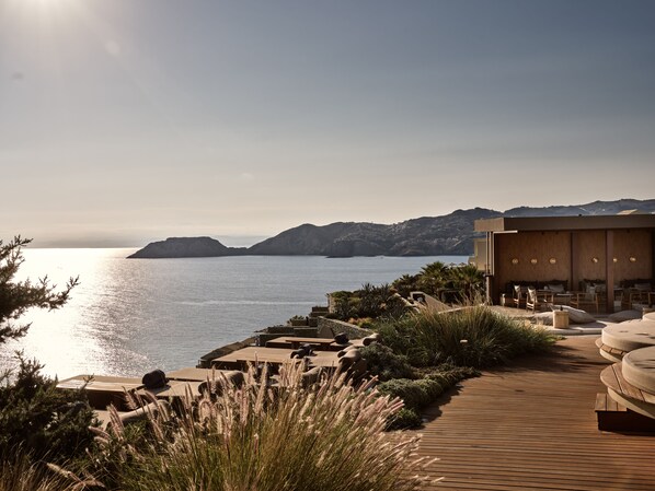A coastal resort with a wooden deck, outdoor seating, and a view of the sea and hills.