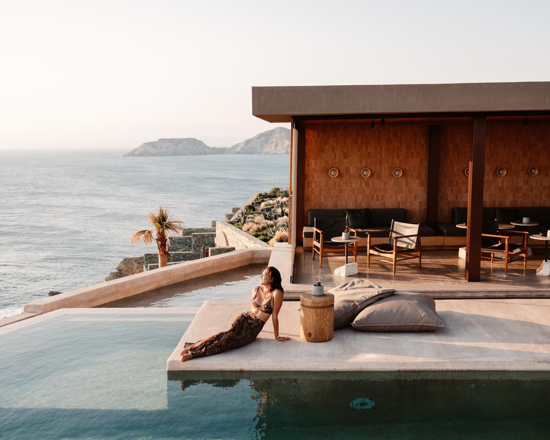 Zona junto a la piscina con vista al mar, una terraza de madera y un espacio de estar con sofá y sillones.