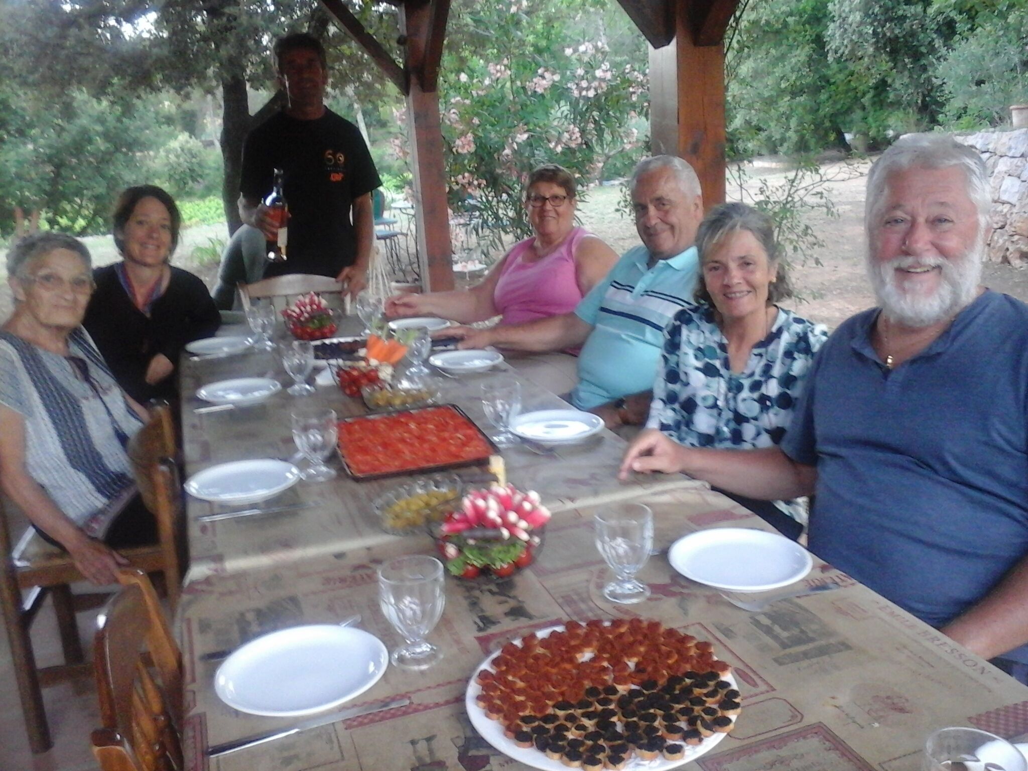 Un groupe de personnes est assis autour d’une table, devant divers plats et boissons, ce qui laisse supposer qu’il s’agit d’un rassemblement ou d’un repas convivial.