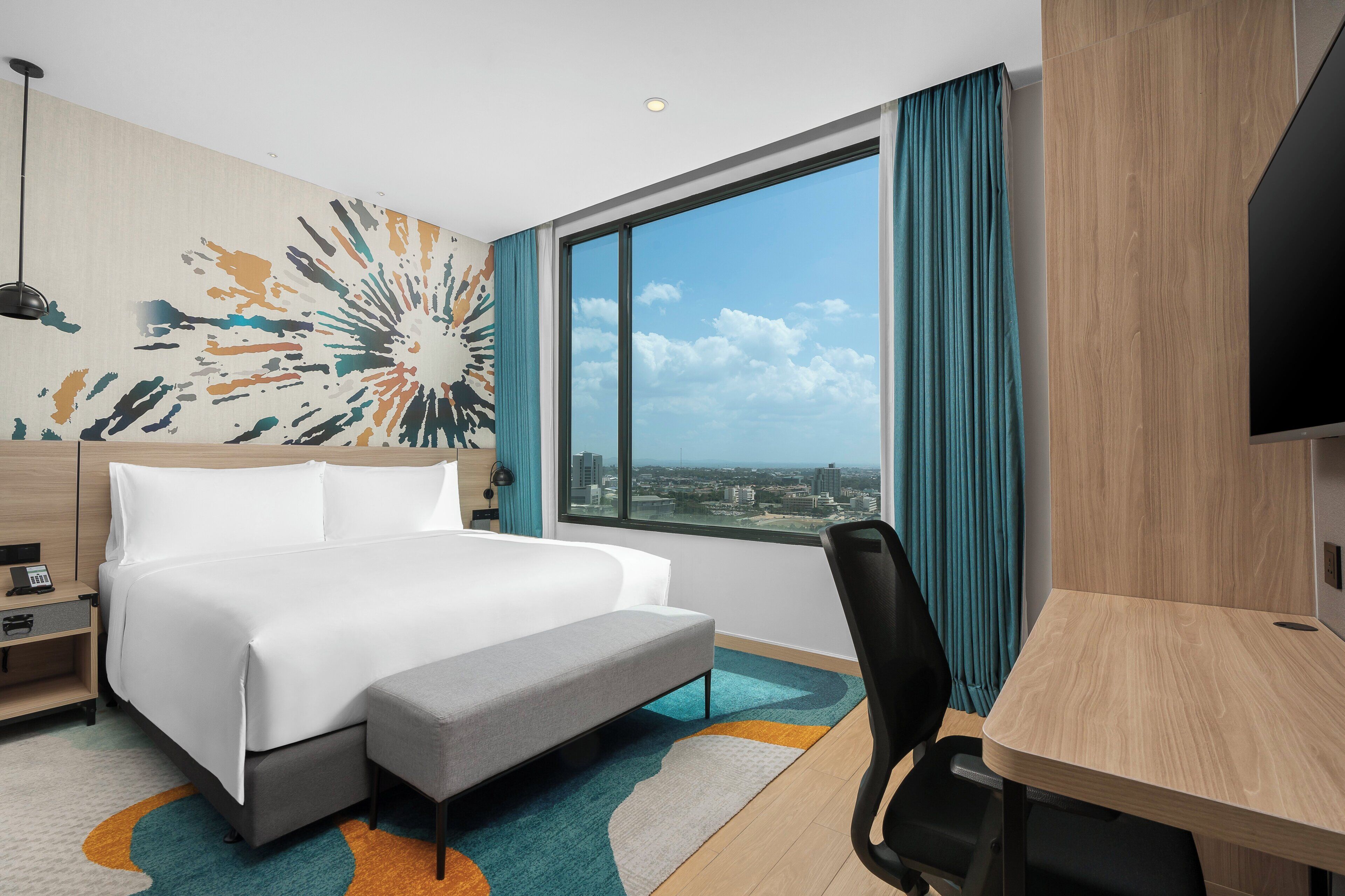 Habitación de hotel con una cama grande, un escritorio, una silla, vistas a la ciudad y un colorido mural abstracto.