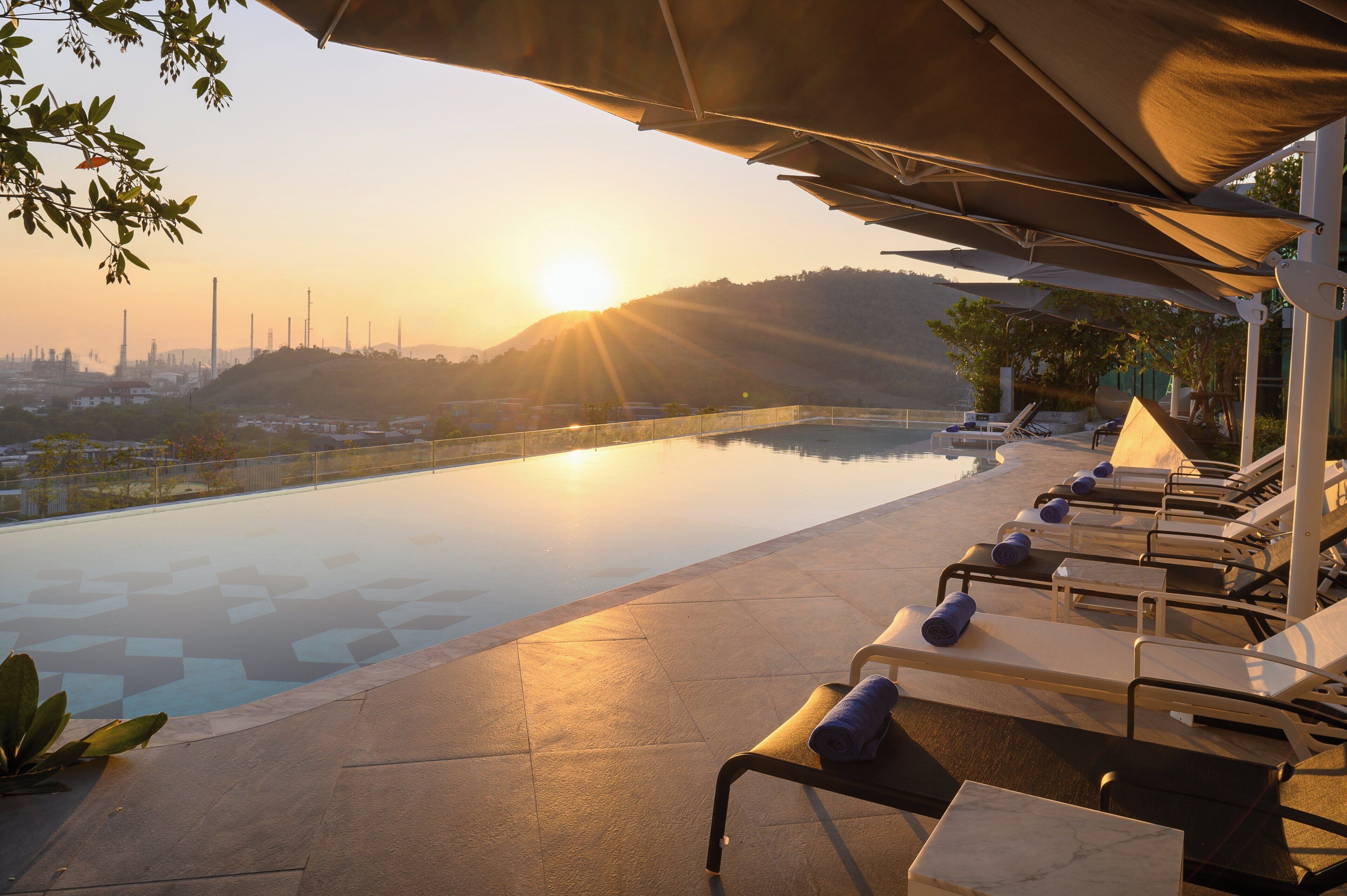 A poolside area with lounge chairs and umbrellas, overlooking a body of water and a distant cityscape during sunset.
