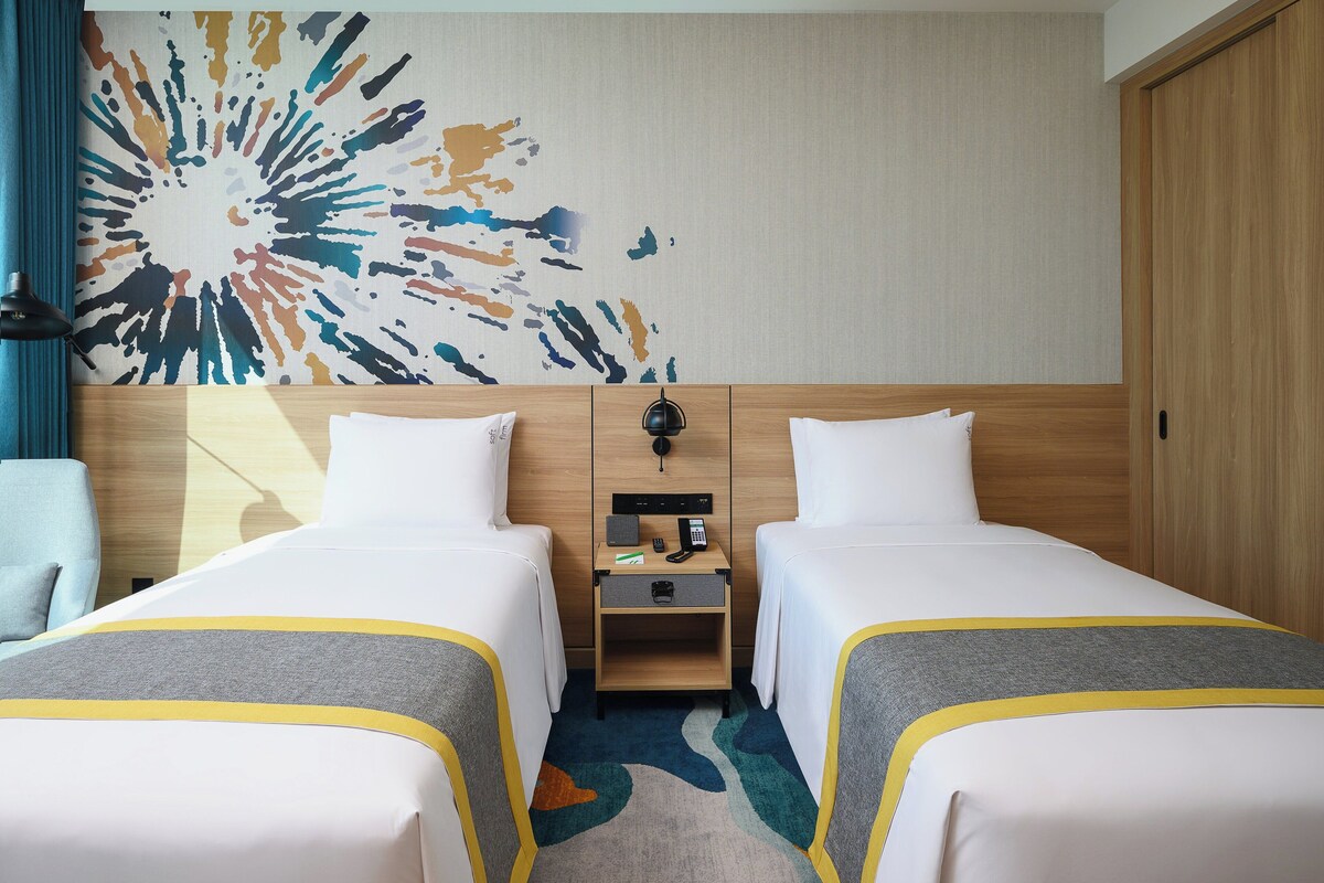 A hotel room with two beds, a wooden headboard, a nightstand with a phone and lamp, and a colorful abstract wall art.