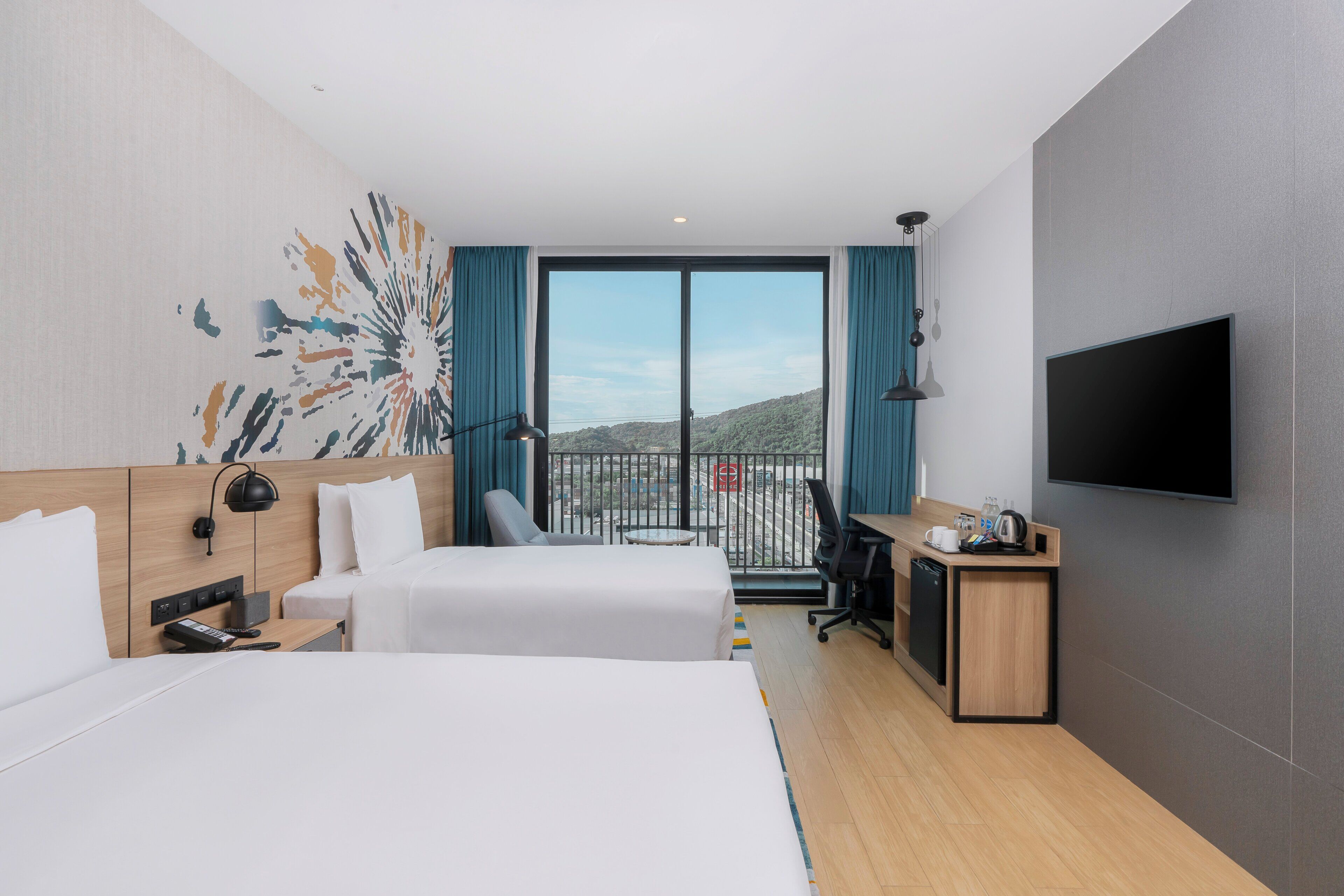 A hotel room with two beds, a desk, a TV, and a balcony with a city view.