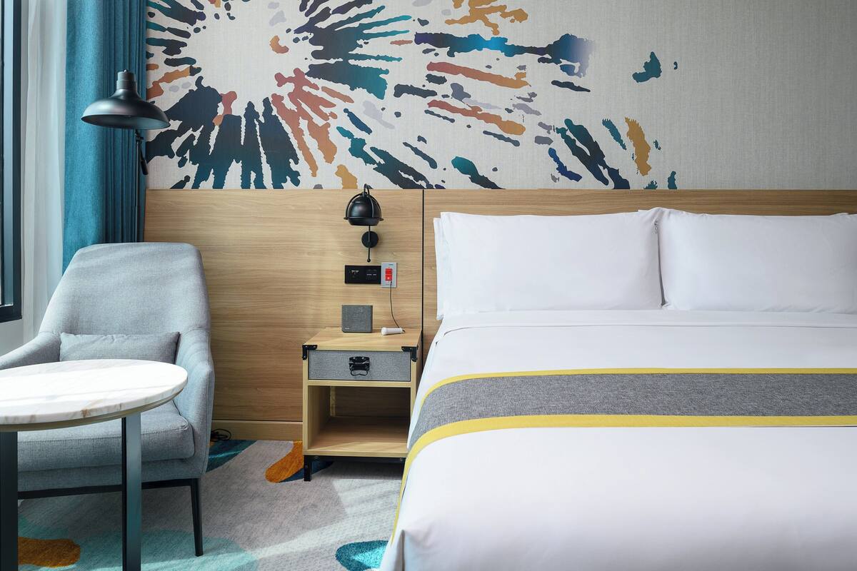 Une chambre d’hôtel comprenant un lit, un bureau, une chaise, une lampe et un mur orné d’un motif abstrait et coloré.
