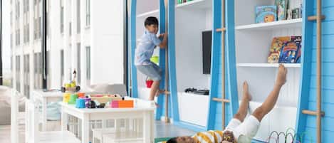 Un niño jugando en una estructura de escalada, otro niño sobre una colchoneta con juguetes, y una estantería con libros y juguetes infantiles.