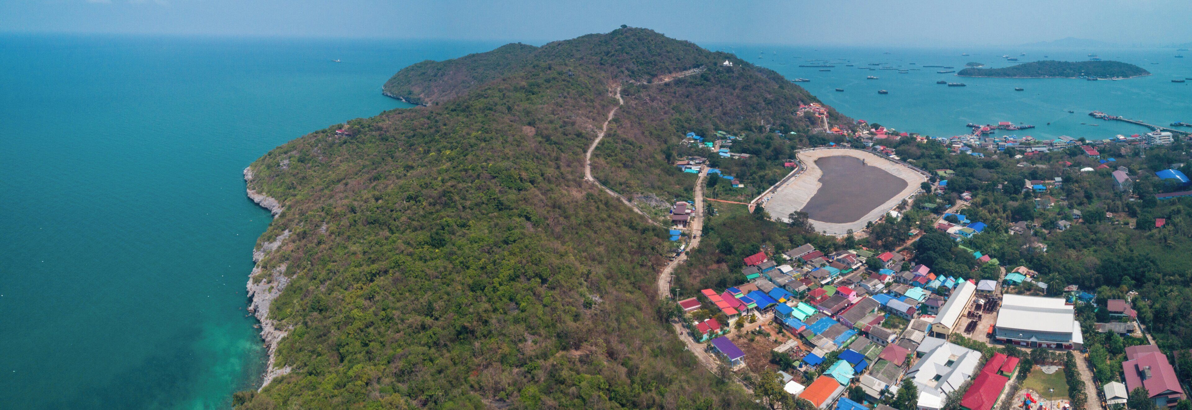 An aerial view of a coastal area with a densely built-up area featuring colorful buildings, a beach, and a forested hill.