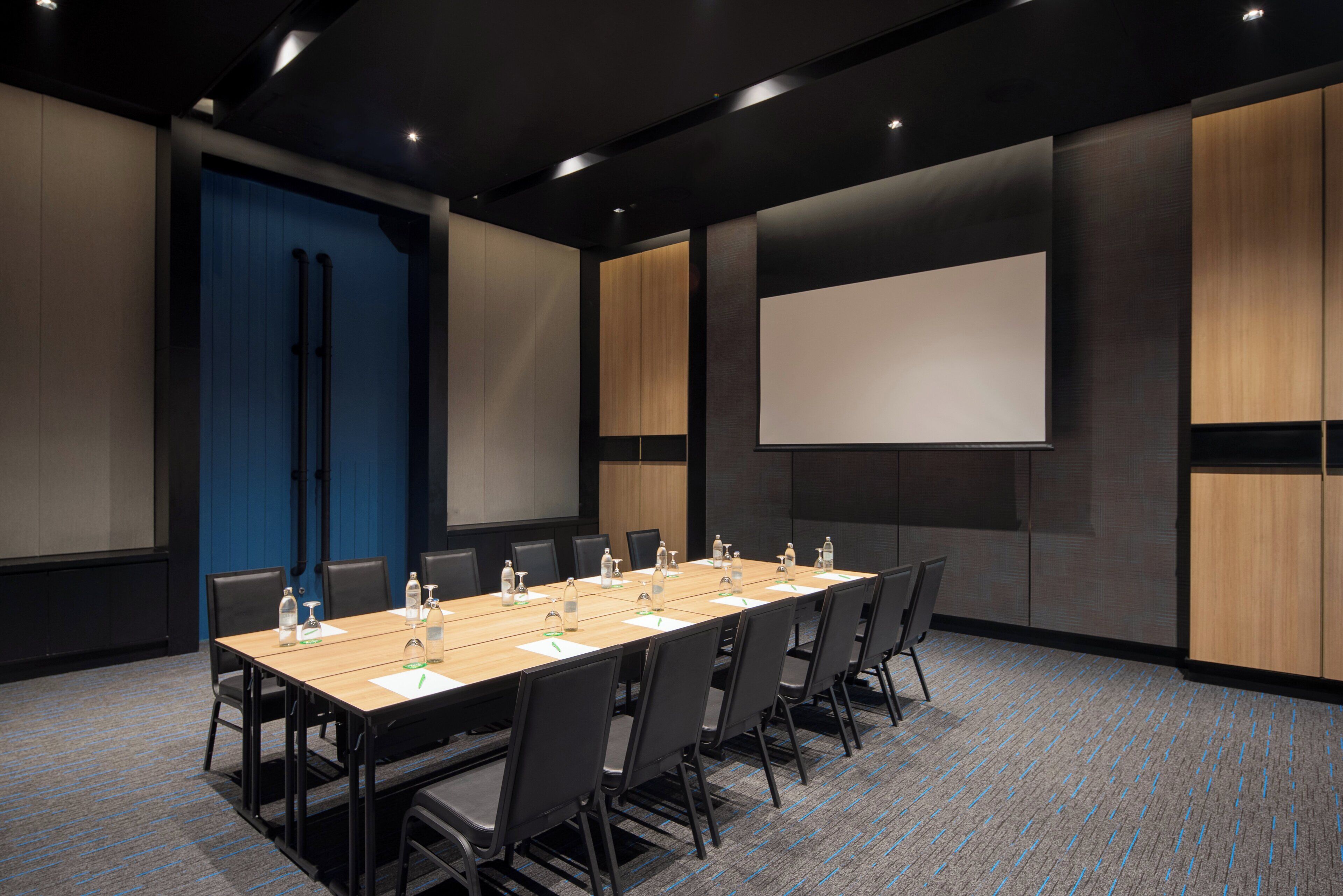 A conference room with a long table, chairs, a projector screen, and a blue door.