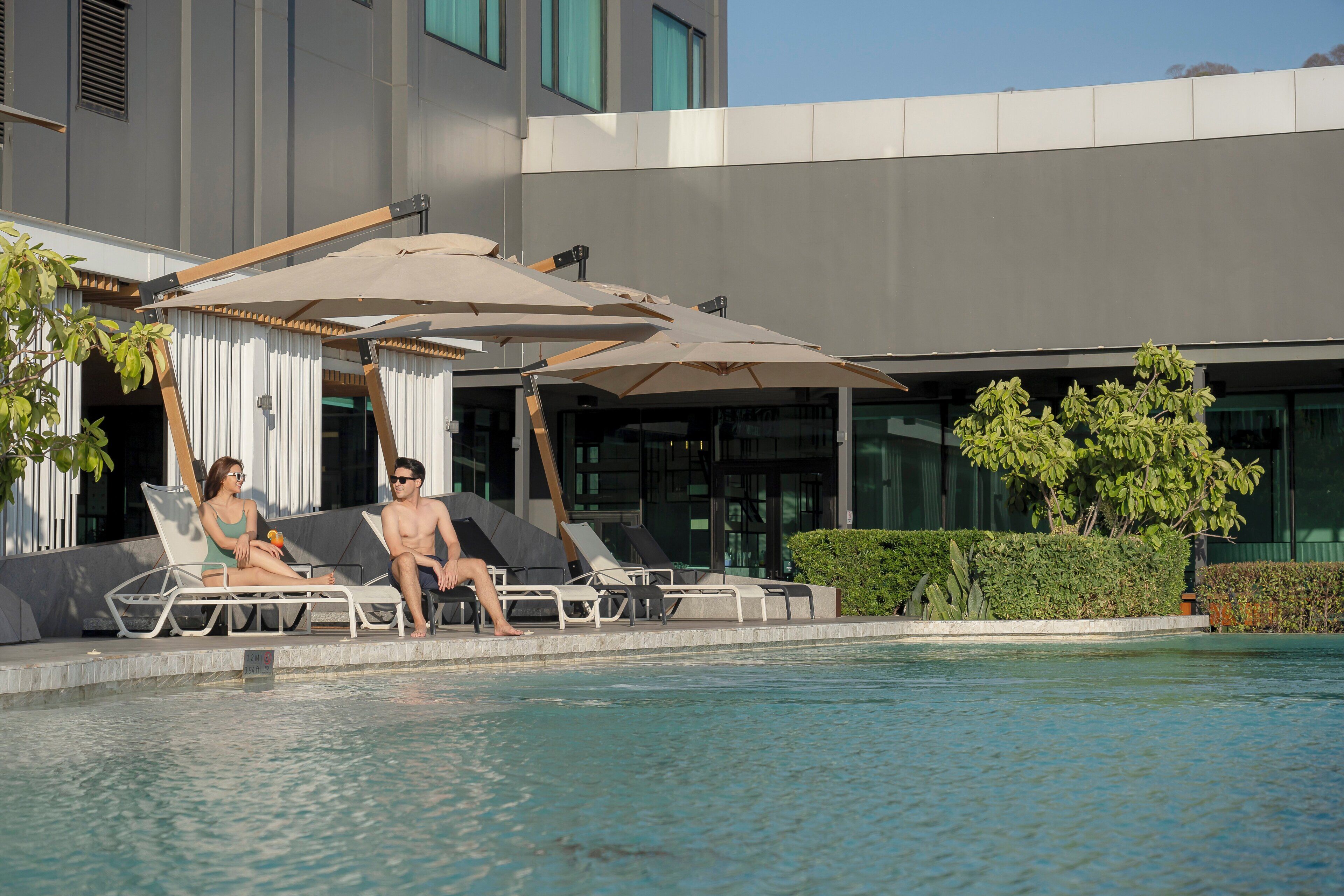A poolside area with lounge chairs and umbrellas, a building with large windows, and a well-maintained hedge.