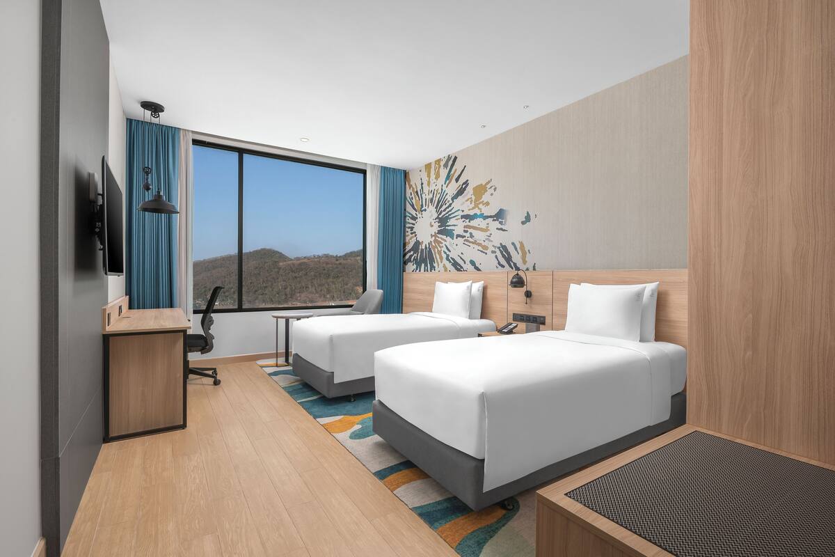 Une chambre d’hôtel avec deux lits, un bureau, une chaise, une télévision et une grande fenêtre donnant sur des collines.