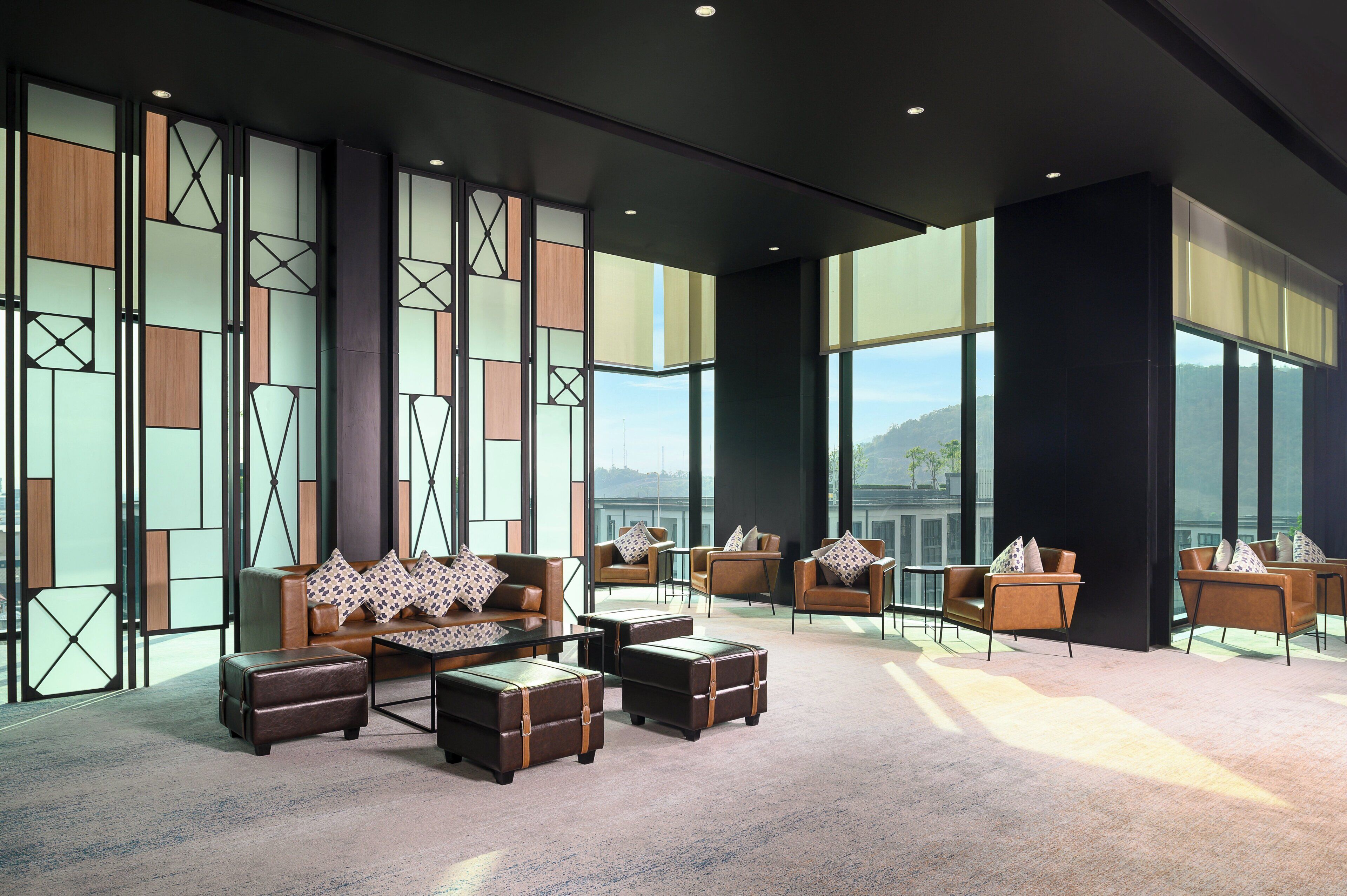 A modern lounge area with leather sofas, armchairs, and ottomans, featuring large windows and a geometric wall design.