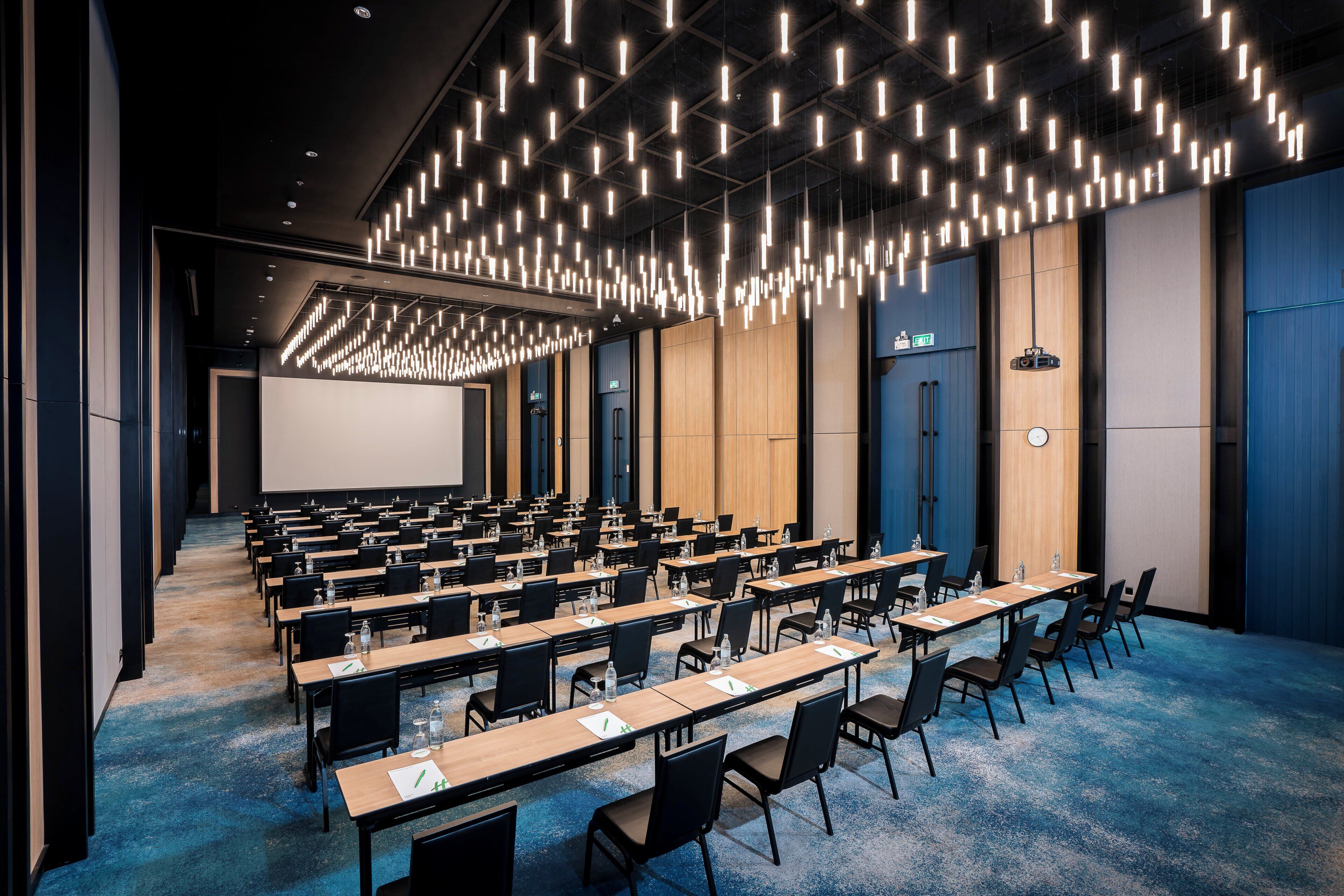 A conference room with rows of tables and chairs, a large screen, and a modern ceiling light fixture.