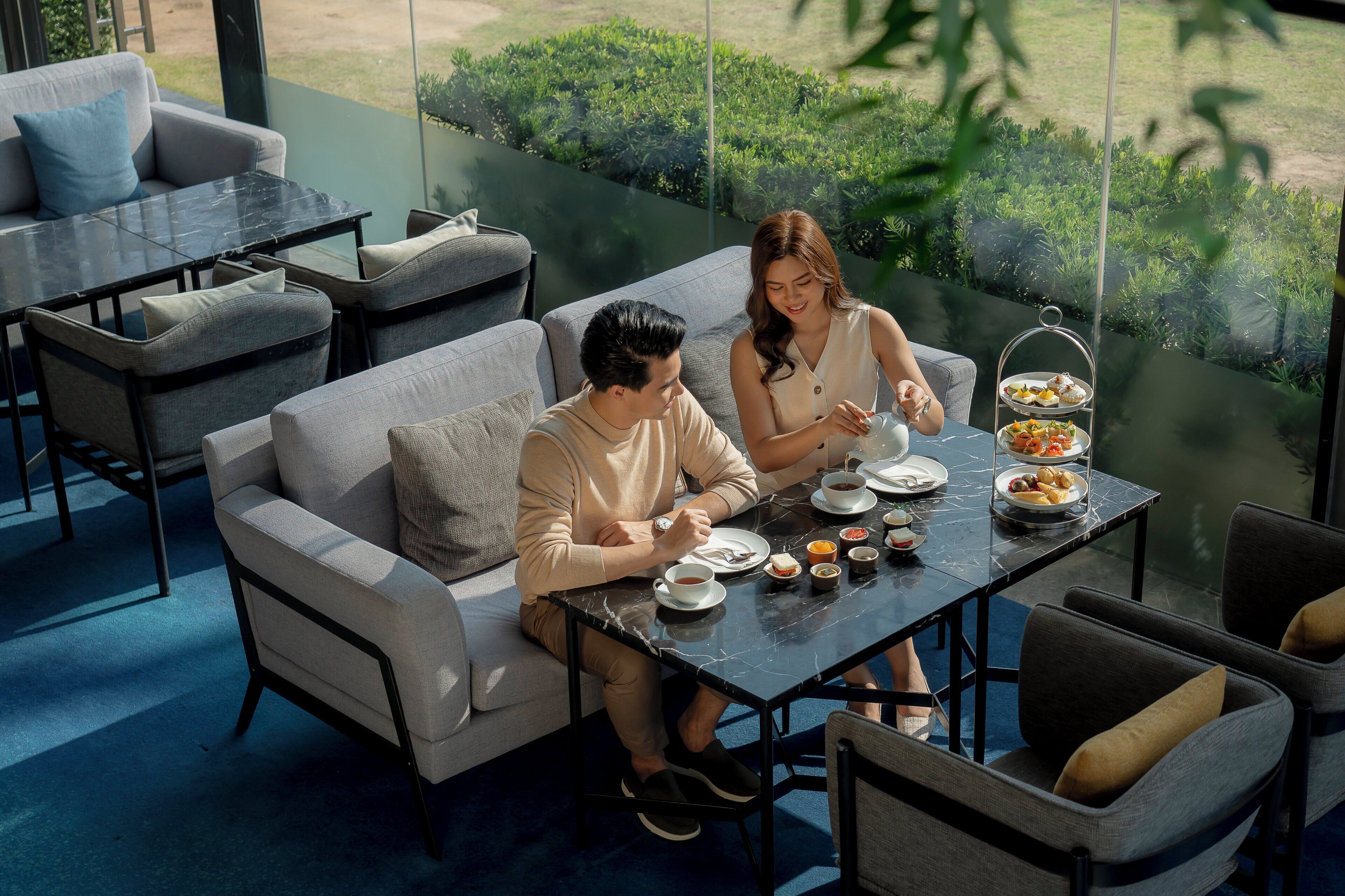 A modern outdoor dining area with a glass partition, cushioned seating, and a table set with food and drinks.