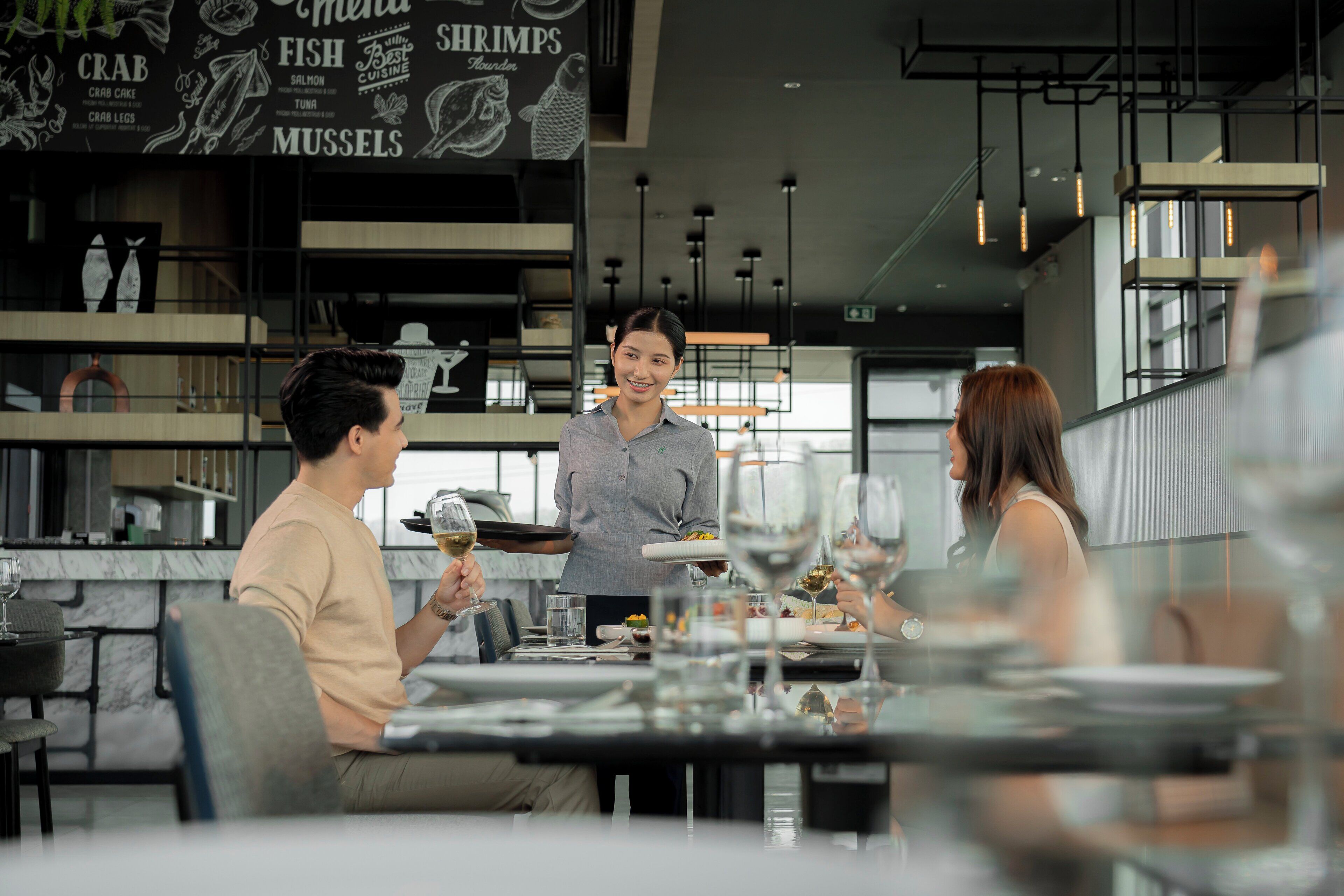 A restaurant interior with a waiter serving a customer, dining tables, and a menu board with seafood options.