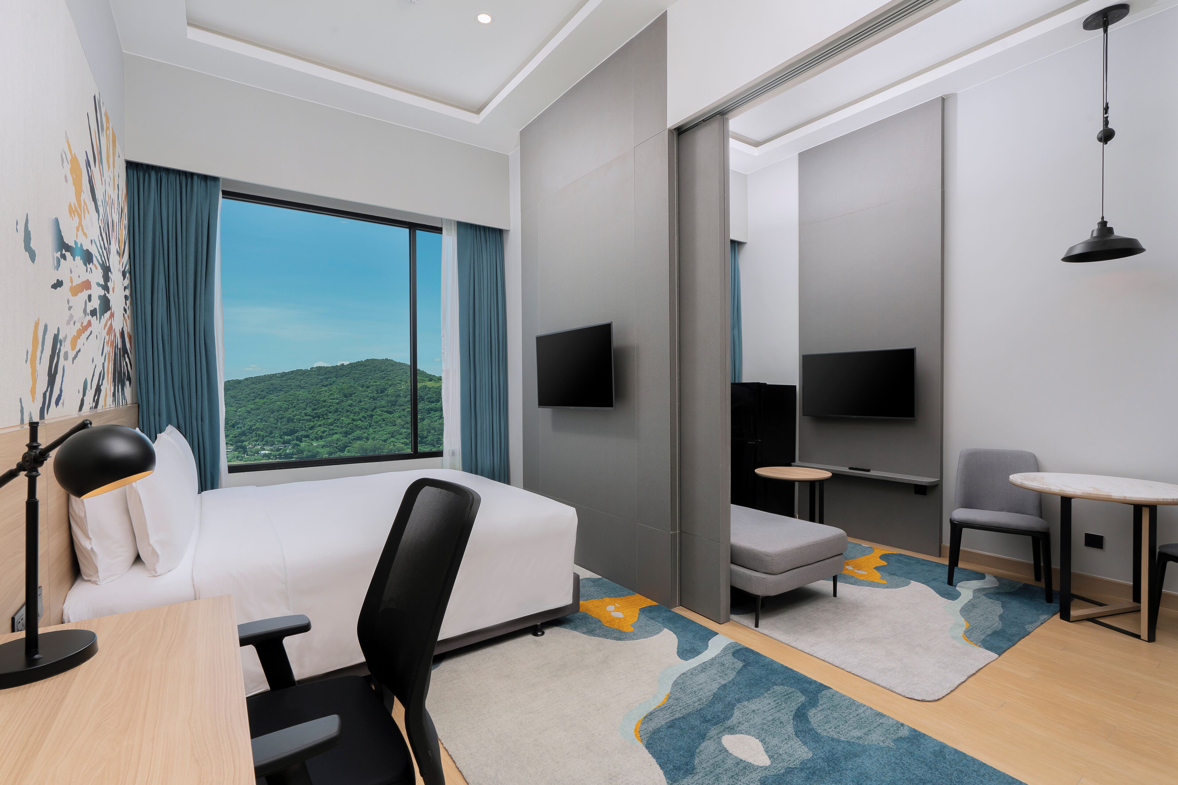 A modern hotel room with a large window, a bed, a desk, a chair, a small table, and a TV.