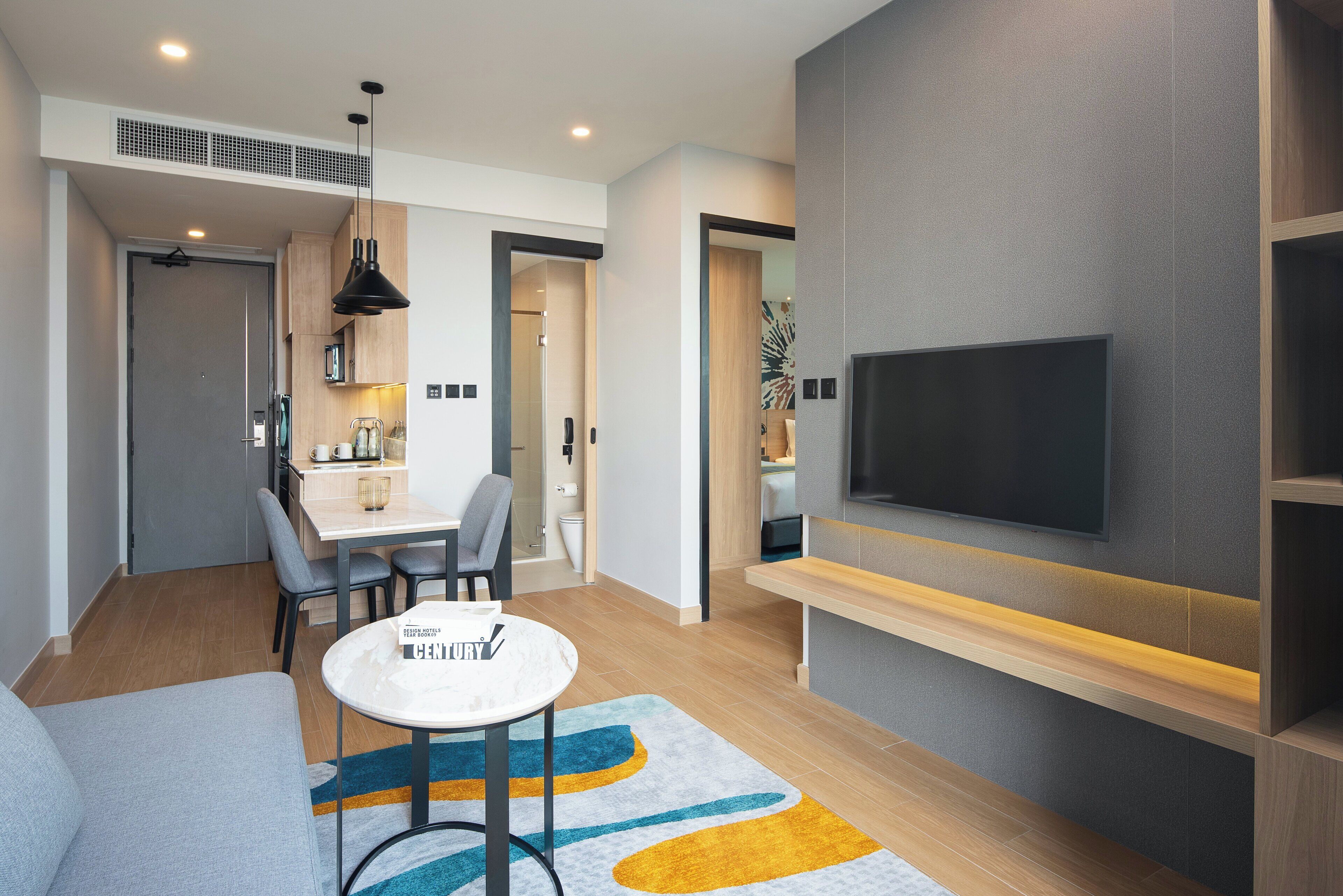A modern living room with a flat-screen TV, a dining area with a table and chairs, and a kitchen visible through an open door.