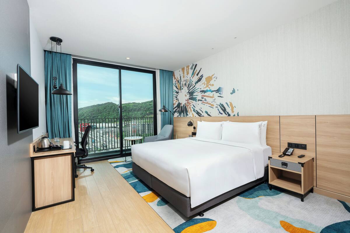 Une chambre d’hôtel avec un grand lit, un bureau doté d’un téléviseur à écran plat, une chaise, une table de chevet avec un téléphone et une vue sur un balcon donnant sur le paysage urbain.