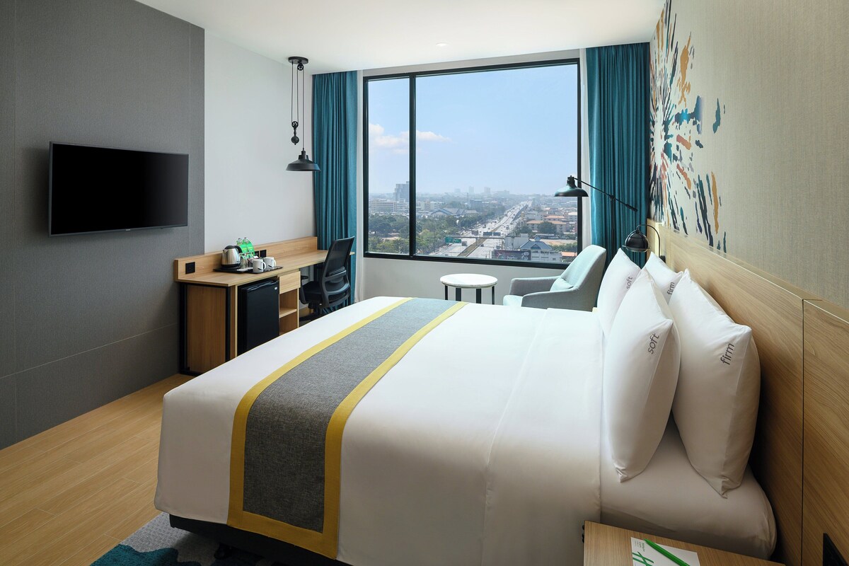 A hotel room with a large bed, a desk, a chair, a TV, and a view of the city.