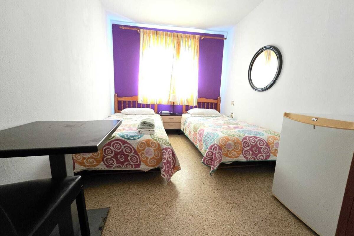 A small, compact room with two single beds, a desk, and a small kitchenette.