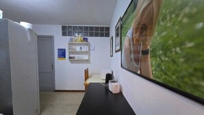 A small, compact room with a kitchenette, a desk, and a chair. There is a refrigerator, a shelf with various items, and a window with a view of greenery. - Low Cost Room Las Llaves 3 (San Bartolomé de Tirajana)