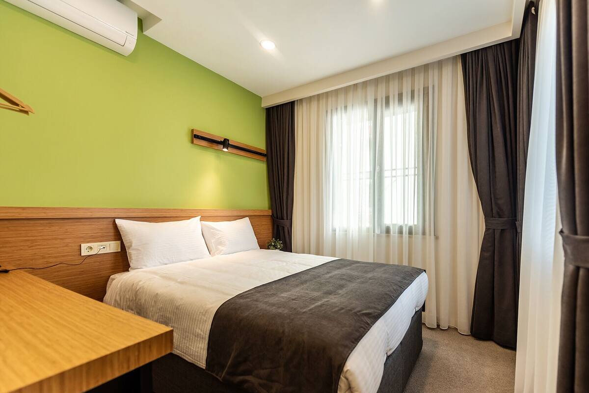 A hotel room with a bed, a wooden desk, a green wall, and a window with curtains.