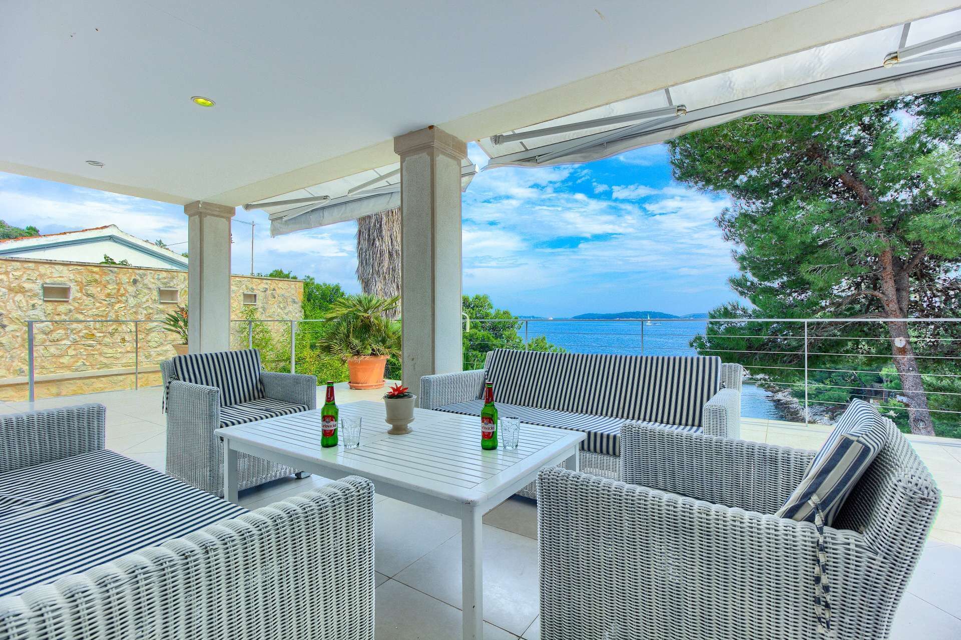 A covered outdoor seating area with wicker furniture, a table, and a view of the sea.